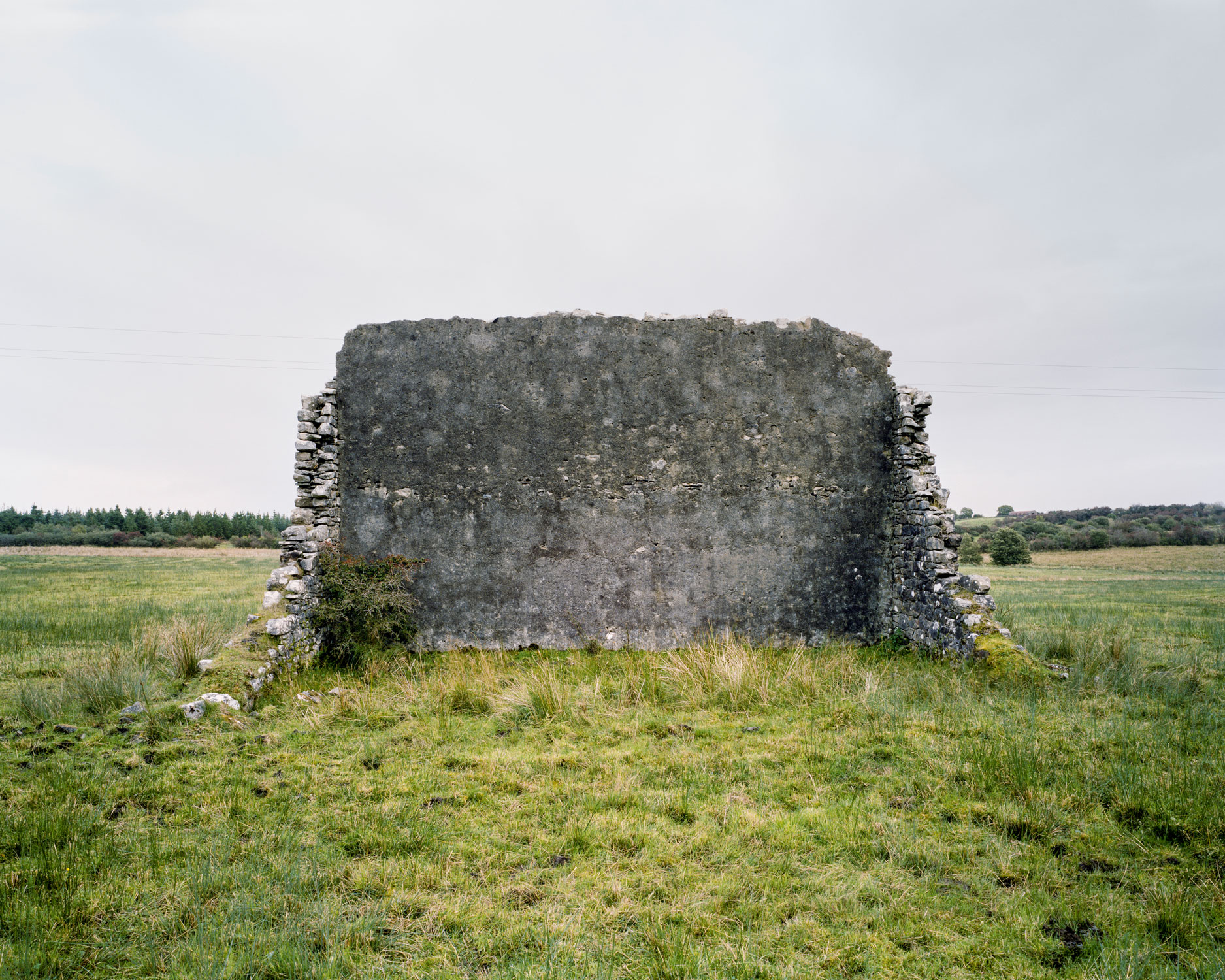 These are relics of a once vibrant chapter of Irish life. Like the dolmens John Montague crafted as a metaphor for the old people of his childhood, the ruins of these abandoned alleys bear silent testimony to a feature of Ireland that has perished through time and modernization. These plain walls were once the thriving open-air theatre's of their day. They were familiar meeting points, where people played games long into long summer evenings, and others gathered for simple companionship and discourse. The alleys came alive at a time in Ireland when human interaction needed little in the way of sophisticated props or accompaniments. In rural Ireland in particular handball captured the public imagination and societies grew around the games and the alleys which staged them. They became prized places of refuge, offering an escape from ordinary, hard-working lives. At the ball alleys, some strategically placed at crossroads in the open countryside, generations happily fraternized. Some were drawn by the magic and noise of a game which has deep Irish roots. But the alley was more than a sporting venue; it became a vital anchor for the community and an invaluable leisure outlet. It also served as an unofficial meeting point for lovers and revolutionaries. Even as a location for executions. It had an open charter - multiple, indiscriminate possibilities. Like Montague's dolmens, some alleys survive - a portion of them intact and a minority enjoying new leases of life. But the majority are disappearing slowly from the landscape to the point where they will eventually become extinct. Many have been merged and camouflaged into other structures, like farmyard sheds or car parks, so that they are barely recognizable or identifiable. Many others are simply abandoned and overgrown. Old handball players and the game's scattered aficionados will recall their last hurrah 50 odd years ago or more, after they had thrived across the country for over a century, before television and better transport began to signal their decline and offer alternative means of entertainment and social collusion - leaving each one in turn to become a tomb for a lost civilization.