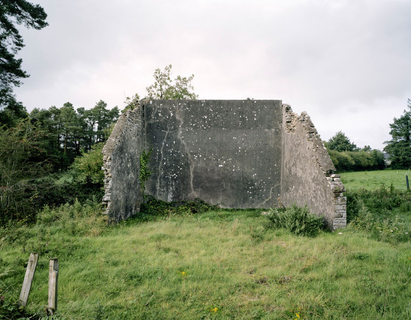These are relics of a once vibrant chapter of Irish life. Like the dolmens John Montague crafted as a metaphor for the old people of his childhood, the ruins of these abandoned alleys bear silent testimony to a feature of Ireland that has perished through time and modernization. These plain walls were once the thriving open-air theatre's of their day. They were familiar meeting points, where people played games long into long summer evenings, and others gathered for simple companionship and discourse. The alleys came alive at a time in Ireland when human interaction needed little in the way of sophisticated props or accompaniments. In rural Ireland in particular handball captured the public imagination and societies grew around the games and the alleys which staged them. They became prized places of refuge, offering an escape from ordinary, hard-working lives. At the ball alleys, some strategically placed at crossroads in the open countryside, generations happily fraternized. Some were drawn by the magic and noise of a game which has deep Irish roots. But the alley was more than a sporting venue; it became a vital anchor for the community and an invaluable leisure outlet. It also served as an unofficial meeting point for lovers and revolutionaries. Even as a location for executions. It had an open charter - multiple, indiscriminate possibilities. Like Montague's dolmens, some alleys survive - a portion of them intact and a minority enjoying new leases of life. But the majority are disappearing slowly from the landscape to the point where they will eventually become extinct. Many have been merged and camouflaged into other structures, like farmyard sheds or car parks, so that they are barely recognizable or identifiable. Many others are simply abandoned and overgrown. Old handball players and the game's scattered aficionados will recall their last hurrah 50 odd years ago or more, after they had thrived across the country for over a century, before television and better transport began to signal their decline and offer alternative means of entertainment and social collusion - leaving each one in turn to become a tomb for a lost civilization.