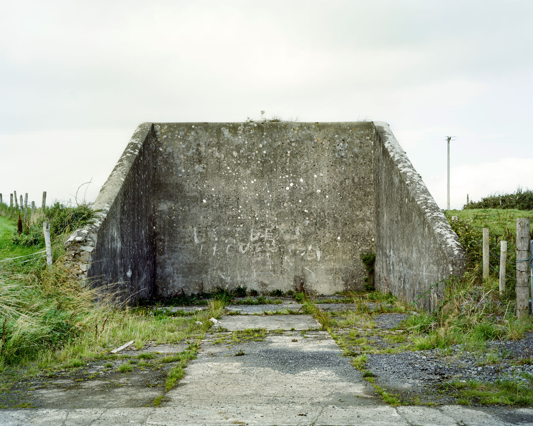 These are relics of a once vibrant chapter of Irish life. Like the dolmens John Montague crafted as a metaphor for the old people of his childhood, the ruins of these abandoned alleys bear silent testimony to a feature of Ireland that has perished through time and modernization. These plain walls were once the thriving open-air theatre's of their day. They were familiar meeting points, where people played games long into long summer evenings, and others gathered for simple companionship and discourse. The alleys came alive at a time in Ireland when human interaction needed little in the way of sophisticated props or accompaniments. In rural Ireland in particular handball captured the public imagination and societies grew around the games and the alleys which staged them. They became prized places of refuge, offering an escape from ordinary, hard-working lives. At the ball alleys, some strategically placed at crossroads in the open countryside, generations happily fraternized. Some were drawn by the magic and noise of a game which has deep Irish roots. But the alley was more than a sporting venue; it became a vital anchor for the community and an invaluable leisure outlet. It also served as an unofficial meeting point for lovers and revolutionaries. Even as a location for executions. It had an open charter - multiple, indiscriminate possibilities. Like Montague's dolmens, some alleys survive - a portion of them intact and a minority enjoying new leases of life. But the majority are disappearing slowly from the landscape to the point where they will eventually become extinct. Many have been merged and camouflaged into other structures, like farmyard sheds or car parks, so that they are barely recognizable or identifiable. Many others are simply abandoned and overgrown. Old handball players and the game's scattered aficionados will recall their last hurrah 50 odd years ago or more, after they had thrived across the country for over a century, before television and better transport began to signal their decline and offer alternative means of entertainment and social collusion - leaving each one in turn to become a tomb for a lost civilization.