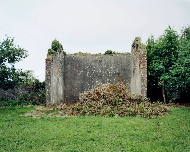 These are relics of a once vibrant chapter of Irish life. Like the dolmens John Montague crafted as a metaphor for the old people of his childhood, the ruins of these abandoned alleys bear silent testimony to a feature of Ireland that has perished through time and modernization. These plain walls were once the thriving open-air theatre's of their day. They were familiar meeting points, where people played games long into long summer evenings, and others gathered for simple companionship and discourse. The alleys came alive at a time in Ireland when human interaction needed little in the way of sophisticated props or accompaniments. In rural Ireland in particular handball captured the public imagination and societies grew around the games and the alleys which staged them. They became prized places of refuge, offering an escape from ordinary, hard-working lives. At the ball alleys, some strategically placed at crossroads in the open countryside, generations happily fraternized. Some were drawn by the magic and noise of a game which has deep Irish roots. But the alley was more than a sporting venue; it became a vital anchor for the community and an invaluable leisure outlet. It also served as an unofficial meeting point for lovers and revolutionaries. Even as a location for executions. It had an open charter - multiple, indiscriminate possibilities. Like Montague's dolmens, some alleys survive - a portion of them intact and a minority enjoying new leases of life. But the majority are disappearing slowly from the landscape to the point where they will eventually become extinct. Many have been merged and camouflaged into other structures, like farmyard sheds or car parks, so that they are barely recognizable or identifiable. Many others are simply abandoned and overgrown. Old handball players and the game's scattered aficionados will recall their last hurrah 50 odd years ago or more, after they had thrived across the country for over a century, before television and better transport began to signal their decline and offer alternative means of entertainment and social collusion - leaving each one in turn to become a tomb for a lost civilization.