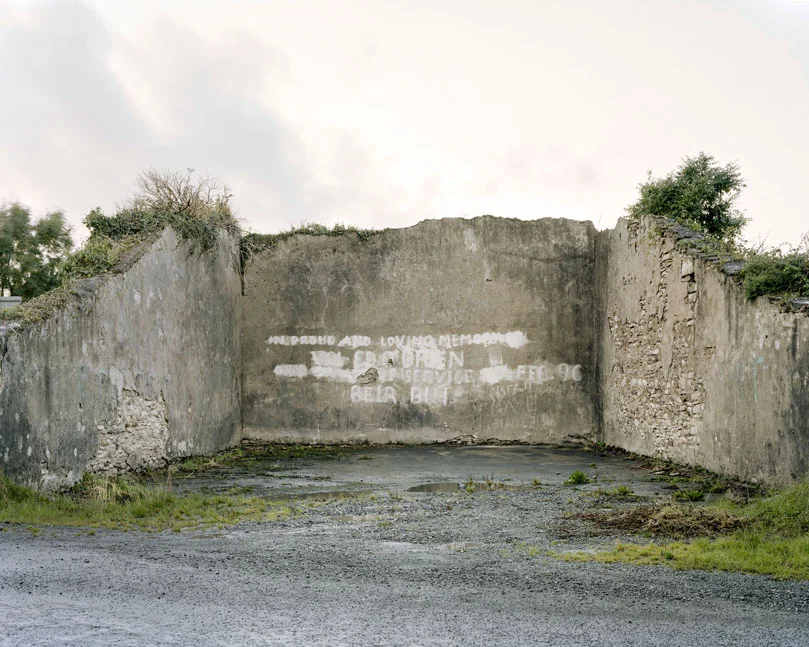 These are relics of a once vibrant chapter of Irish life. Like the dolmens John Montague crafted as a metaphor for the old people of his childhood, the ruins of these abandoned alleys bear silent testimony to a feature of Ireland that has perished through time and modernization. These plain walls were once the thriving open-air theatre's of their day. They were familiar meeting points, where people played games long into long summer evenings, and others gathered for simple companionship and discourse. The alleys came alive at a time in Ireland when human interaction needed little in the way of sophisticated props or accompaniments. In rural Ireland in particular handball captured the public imagination and societies grew around the games and the alleys which staged them. They became prized places of refuge, offering an escape from ordinary, hard-working lives. At the ball alleys, some strategically placed at crossroads in the open countryside, generations happily fraternized. Some were drawn by the magic and noise of a game which has deep Irish roots. But the alley was more than a sporting venue; it became a vital anchor for the community and an invaluable leisure outlet. It also served as an unofficial meeting point for lovers and revolutionaries. Even as a location for executions. It had an open charter - multiple, indiscriminate possibilities. Like Montague's dolmens, some alleys survive - a portion of them intact and a minority enjoying new leases of life. But the majority are disappearing slowly from the landscape to the point where they will eventually become extinct. Many have been merged and camouflaged into other structures, like farmyard sheds or car parks, so that they are barely recognizable or identifiable. Many others are simply abandoned and overgrown. Old handball players and the game's scattered aficionados will recall their last hurrah 50 odd years ago or more, after they had thrived across the country for over a century, before television and better transport began to signal their decline and offer alternative means of entertainment and social collusion - leaving each one in turn to become a tomb for a lost civilization.