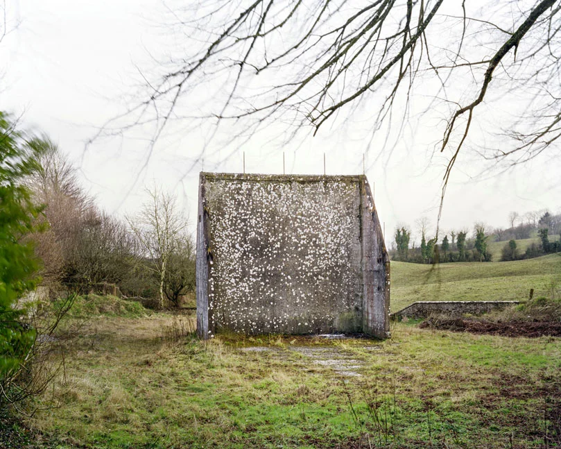 These are relics of a once vibrant chapter of Irish life. Like the dolmens John Montague crafted as a metaphor for the old people of his childhood, the ruins of these abandoned alleys bear silent testimony to a feature of Ireland that has perished through time and modernization. These plain walls were once the thriving open-air theatre's of their day. They were familiar meeting points, where people played games long into long summer evenings, and others gathered for simple companionship and discourse. The alleys came alive at a time in Ireland when human interaction needed little in the way of sophisticated props or accompaniments. In rural Ireland in particular handball captured the public imagination and societies grew around the games and the alleys which staged them. They became prized places of refuge, offering an escape from ordinary, hard-working lives. At the ball alleys, some strategically placed at crossroads in the open countryside, generations happily fraternized. Some were drawn by the magic and noise of a game which has deep Irish roots. But the alley was more than a sporting venue; it became a vital anchor for the community and an invaluable leisure outlet. It also served as an unofficial meeting point for lovers and revolutionaries. Even as a location for executions. It had an open charter - multiple, indiscriminate possibilities. Like Montague's dolmens, some alleys survive - a portion of them intact and a minority enjoying new leases of life. But the majority are disappearing slowly from the landscape to the point where they will eventually become extinct. Many have been merged and camouflaged into other structures, like farmyard sheds or car parks, so that they are barely recognizable or identifiable. Many others are simply abandoned and overgrown. Old handball players and the game's scattered aficionados will recall their last hurrah 50 odd years ago or more, after they had thrived across the country for over a century, before television and better transport began to signal their decline and offer alternative means of entertainment and social collusion - leaving each one in turn to become a tomb for a lost civilization.
