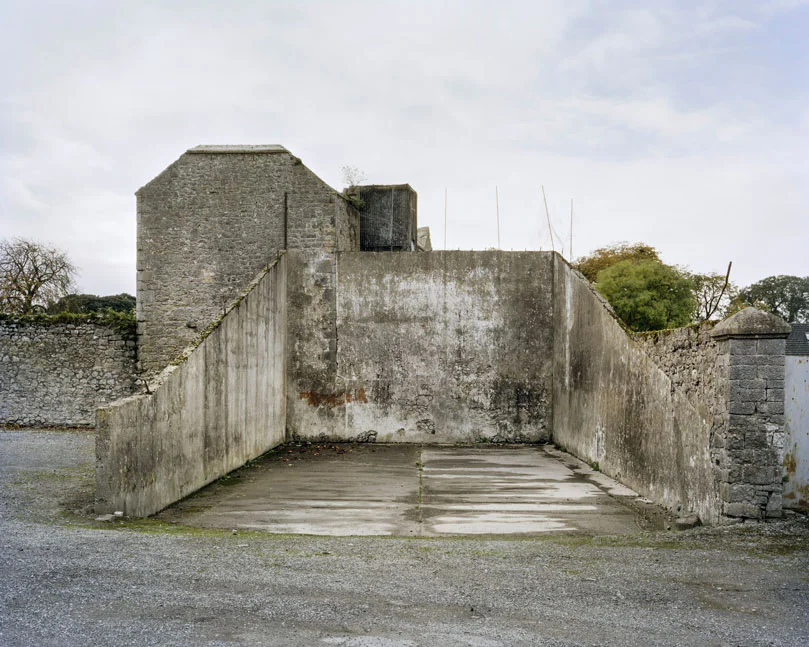 These are relics of a once vibrant chapter of Irish life. Like the dolmens John Montague crafted as a metaphor for the old people of his childhood, the ruins of these abandoned alleys bear silent testimony to a feature of Ireland that has perished through time and modernization. These plain walls were once the thriving open-air theatre's of their day. They were familiar meeting points, where people played games long into long summer evenings, and others gathered for simple companionship and discourse. The alleys came alive at a time in Ireland when human interaction needed little in the way of sophisticated props or accompaniments. In rural Ireland in particular handball captured the public imagination and societies grew around the games and the alleys which staged them. They became prized places of refuge, offering an escape from ordinary, hard-working lives. At the ball alleys, some strategically placed at crossroads in the open countryside, generations happily fraternized. Some were drawn by the magic and noise of a game which has deep Irish roots. But the alley was more than a sporting venue; it became a vital anchor for the community and an invaluable leisure outlet. It also served as an unofficial meeting point for lovers and revolutionaries. Even as a location for executions. It had an open charter - multiple, indiscriminate possibilities. Like Montague's dolmens, some alleys survive - a portion of them intact and a minority enjoying new leases of life. But the majority are disappearing slowly from the landscape to the point where they will eventually become extinct. Many have been merged and camouflaged into other structures, like farmyard sheds or car parks, so that they are barely recognizable or identifiable. Many others are simply abandoned and overgrown. Old handball players and the game's scattered aficionados will recall their last hurrah 50 odd years ago or more, after they had thrived across the country for over a century, before television and better transport began to signal their decline and offer alternative means of entertainment and social collusion - leaving each one in turn to become a tomb for a lost civilization.