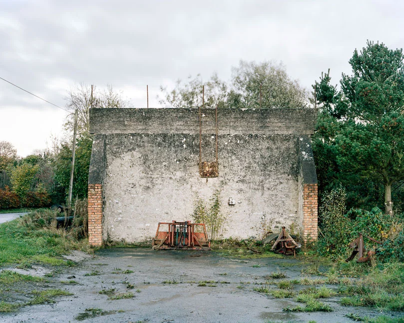 These are relics of a once vibrant chapter of Irish life. Like the dolmens John Montague crafted as a metaphor for the old people of his childhood, the ruins of these abandoned alleys bear silent testimony to a feature of Ireland that has perished through time and modernization. These plain walls were once the thriving open-air theatre's of their day. They were familiar meeting points, where people played games long into long summer evenings, and others gathered for simple companionship and discourse. The alleys came alive at a time in Ireland when human interaction needed little in the way of sophisticated props or accompaniments. In rural Ireland in particular handball captured the public imagination and societies grew around the games and the alleys which staged them. They became prized places of refuge, offering an escape from ordinary, hard-working lives. At the ball alleys, some strategically placed at crossroads in the open countryside, generations happily fraternized. Some were drawn by the magic and noise of a game which has deep Irish roots. But the alley was more than a sporting venue; it became a vital anchor for the community and an invaluable leisure outlet. It also served as an unofficial meeting point for lovers and revolutionaries. Even as a location for executions. It had an open charter - multiple, indiscriminate possibilities. Like Montague's dolmens, some alleys survive - a portion of them intact and a minority enjoying new leases of life. But the majority are disappearing slowly from the landscape to the point where they will eventually become extinct. Many have been merged and camouflaged into other structures, like farmyard sheds or car parks, so that they are barely recognizable or identifiable. Many others are simply abandoned and overgrown. Old handball players and the game's scattered aficionados will recall their last hurrah 50 odd years ago or more, after they had thrived across the country for over a century, before television and better transport began to signal their decline and offer alternative means of entertainment and social collusion - leaving each one in turn to become a tomb for a lost civilization.