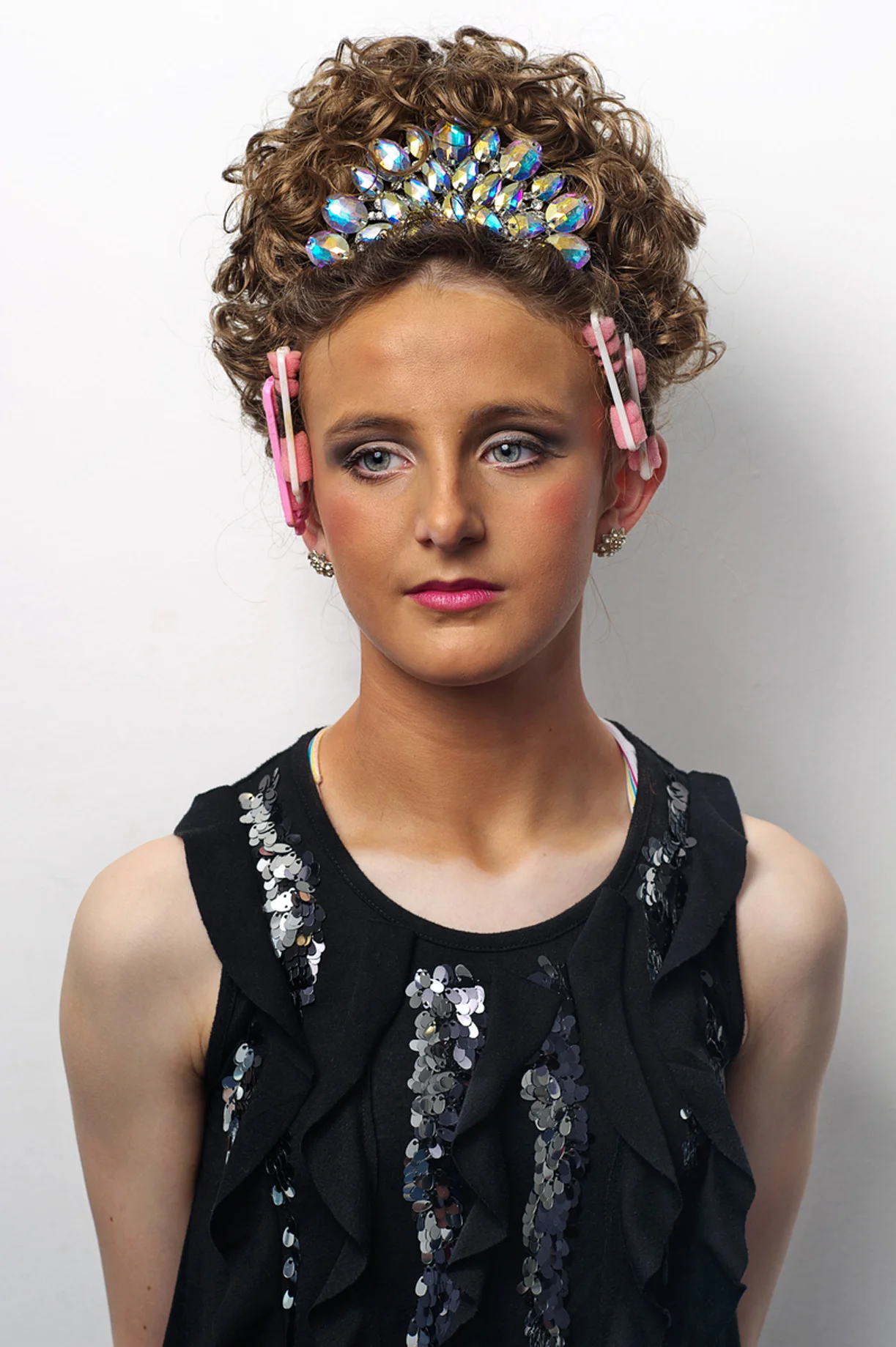 In 1994 during a seven-minute interval at the Eurovision Song Contest in Dublin a group of dancers took to the stage and proceeded to blow the audience away, including millions on television. Fronted by Michael Flatley and Jean Butler, soon to be household names, and accompanied by vibrant and relentless traditionalIrish music, this was the first public performance of the global phenomenon that would become known as Riverdance. Now fast forward to Belfast in April, 2012. Up to 4,500 competitors took part in the Irish World Dance Championships, an event that began in more humble circumstances in Dublin in 1970 – its aim being to promote Gaelic dance culture. Since then its popularity has gone through the roof with contestants travelling from all over the world to compete. The show is more than a dance discipline in the strict sense; the ‘look’ is also deemed important and almost an industry it itself. Contestants spray on fake-tan, carefully apply make up, attach wigs and tiaras and sport dresses that in some cases cost up to 4,000 Euro. It is an expensive environment as well as being a highly charged and extremely competitive one. Why do they spend so much time and money on costume? To catch the judges’ attention is the common explanation. Critics claim it is over-indulgent and garish and has little to do with the reason they are there – to dance, to promote culture. Advocates insist the dance is the most important facet on which contestants are scored and that irrespective of how ornate one’s embroidered dress, these choices are entirely discretionary. There are no hard rules governing dress sense. But it is widely accepted by those who compete and their families that unless you stand out visually then there is a danger you may be ignored. 