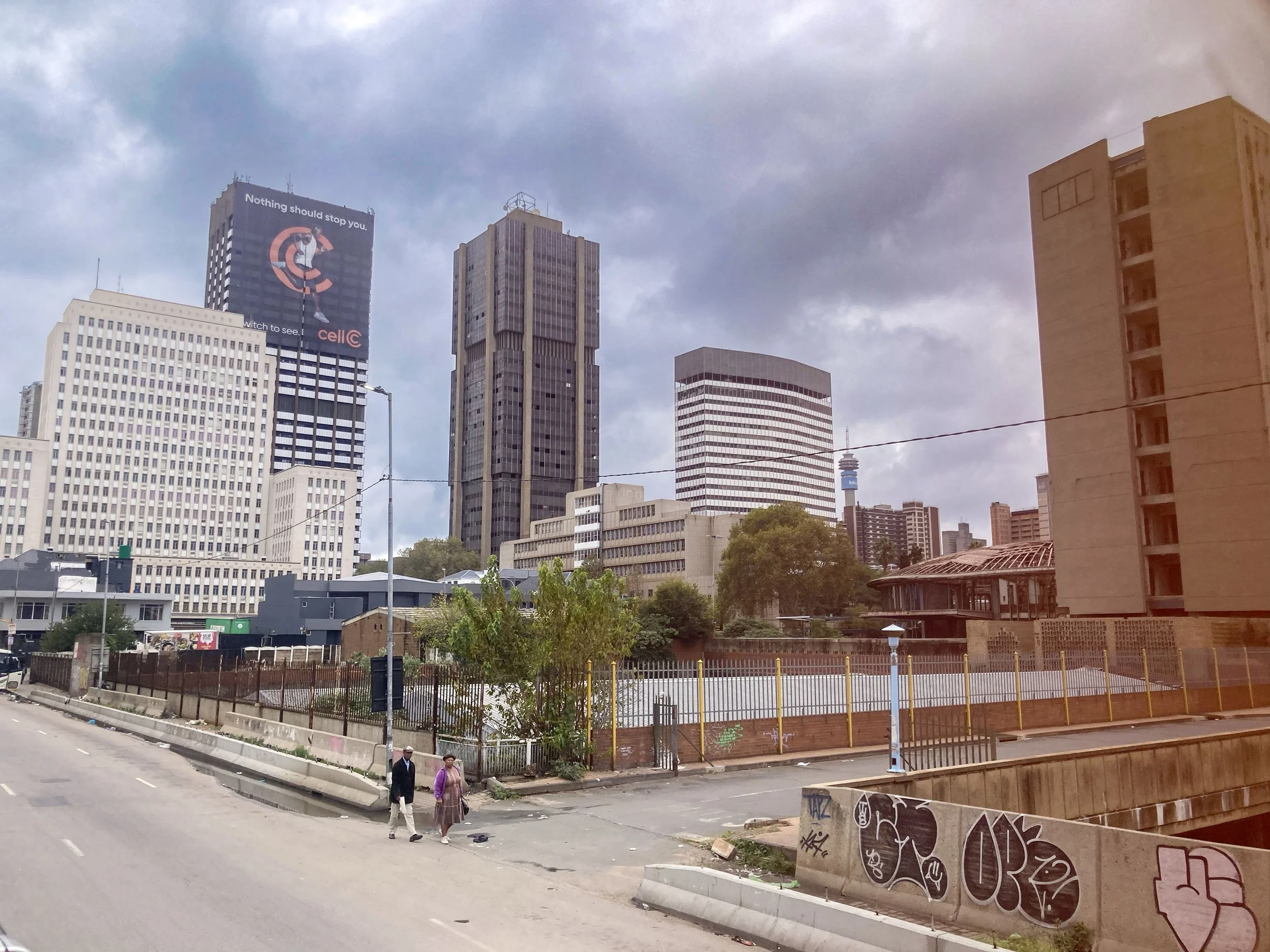 Vue de la skyline urbaine avec plusieurs gratte-ciel, un panneau d'affichage publicitaire, des graffitis sur un mur, une rue avec deux personnes marchant, un ciel nuageux.