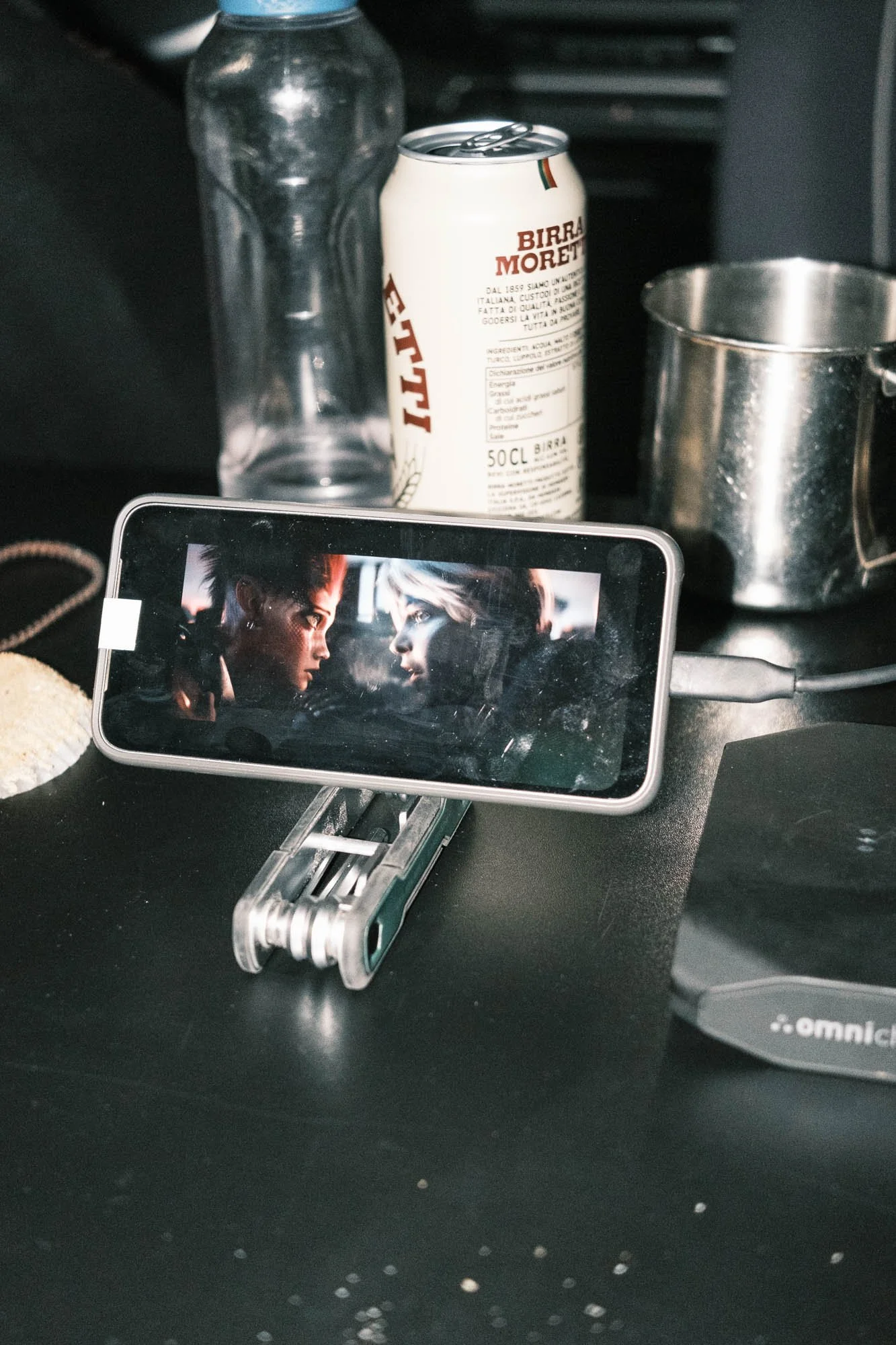 Une canette de bière posé sur une table, une bouteille d'eau, un verre en métal, un téléphone affichant deux personnages face à face, une souris d'ordinateur et une boîte noire à côté.