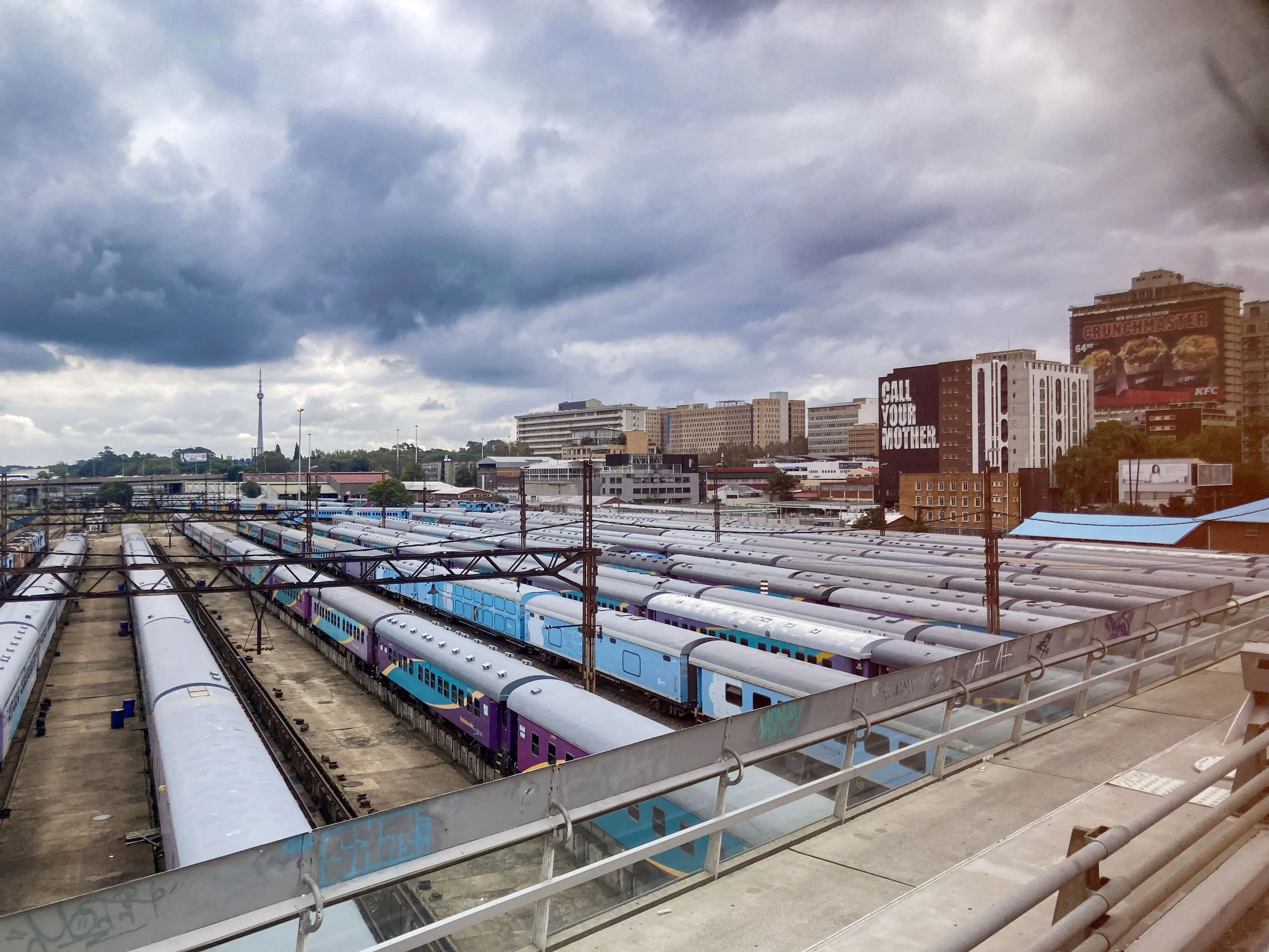 Vue d'une gare de trains avec plusieurs locomotives colorées et des trains stationnés sous un ciel nuageux, dans une ville urbaine.