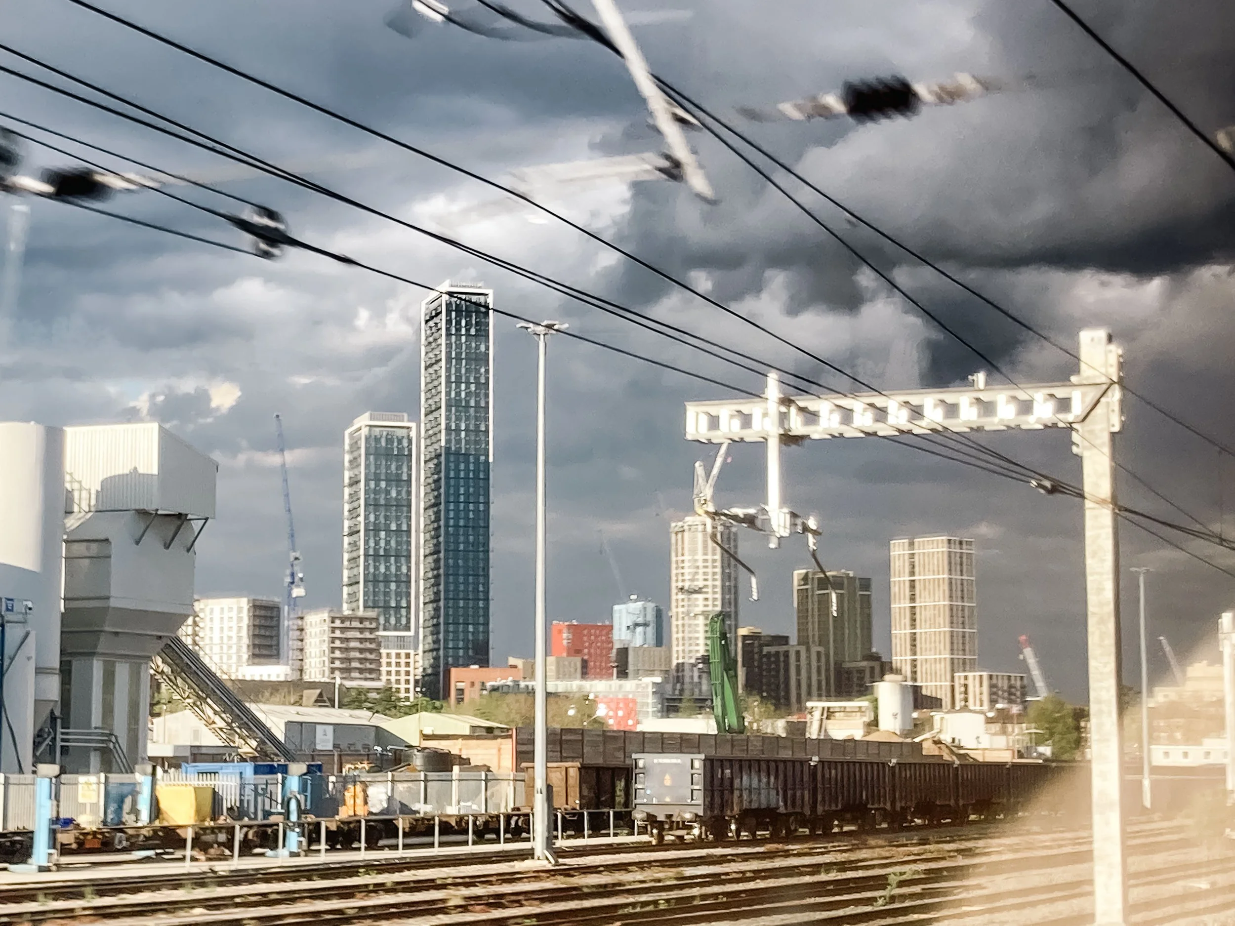 Vue d'une ville avec des gratte-ciel modernes sous un ciel nuageux, vue depuis un train avec des rails en premier plan et des câbles électriques traversant l'image.