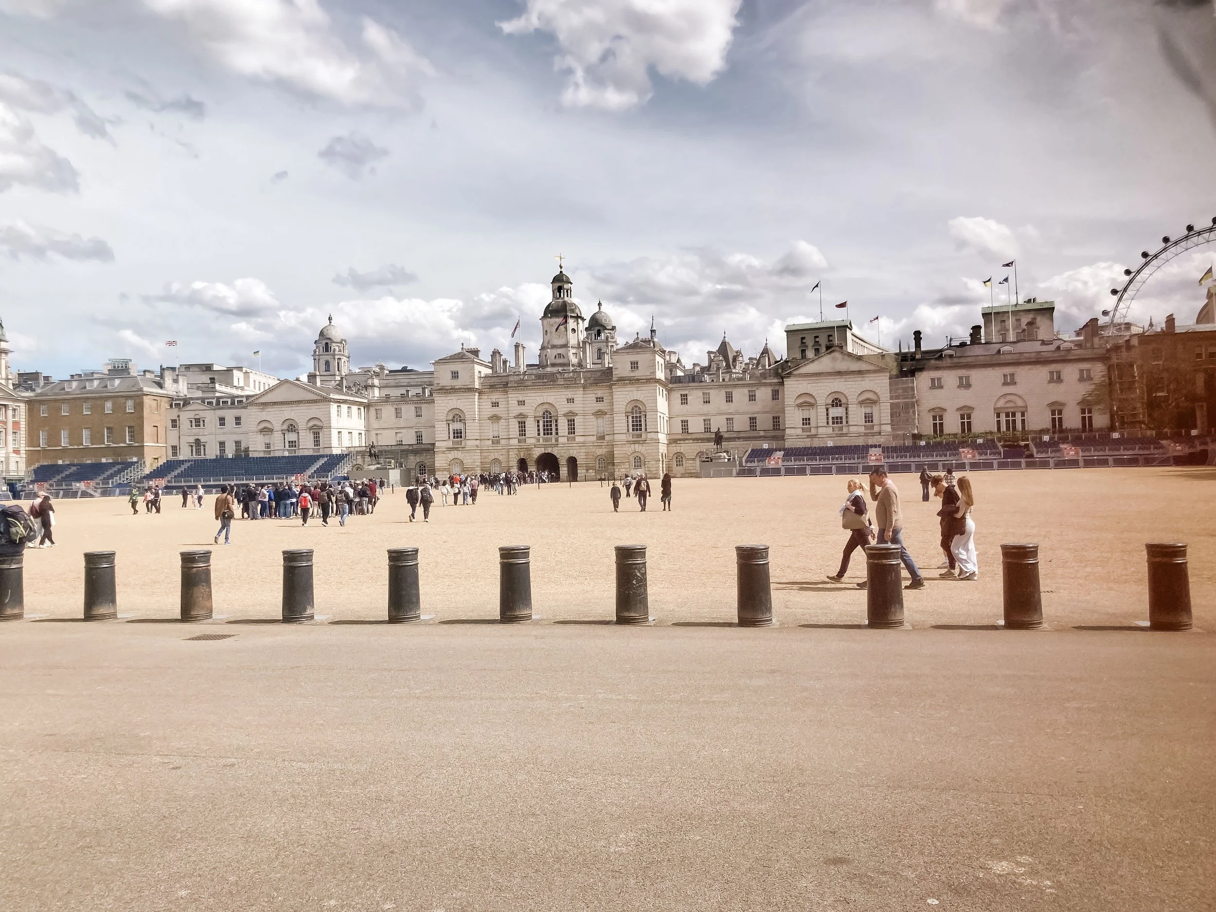 Vue d'un grand bâtiment historique avec plusieurs tours et dômes au centre d'une place, entourée de murs en pierre, avec des gens qui marchent et des barrières en métal devant le bâtiment, sous un ciel partiellement nuageux.