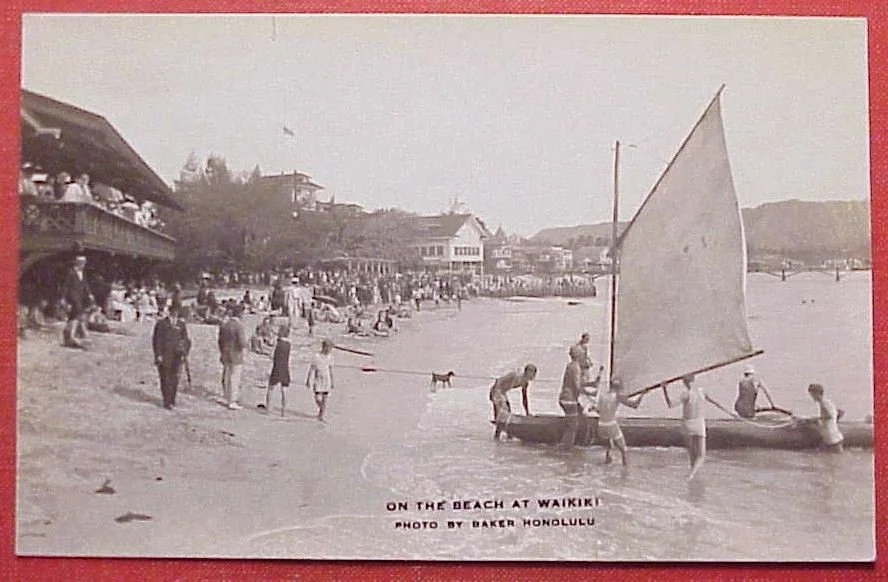  1919 photographic postcard featuring an outrigger with sprit sail on the beach near what looks the Outrigger Canoe Clubhouse 