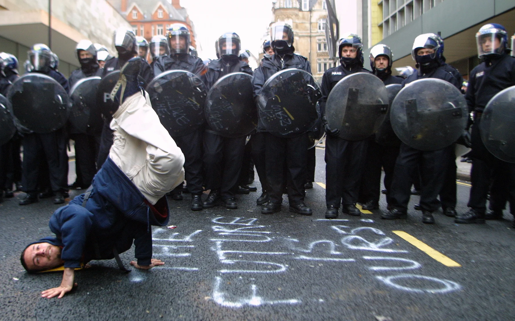 A protester busting a move in front of a line of Riot Police, With Graffiti, 'The Future Is Ours' Written on the road, Mayday Riots, London 2001 © Lee Jones / PYMCA