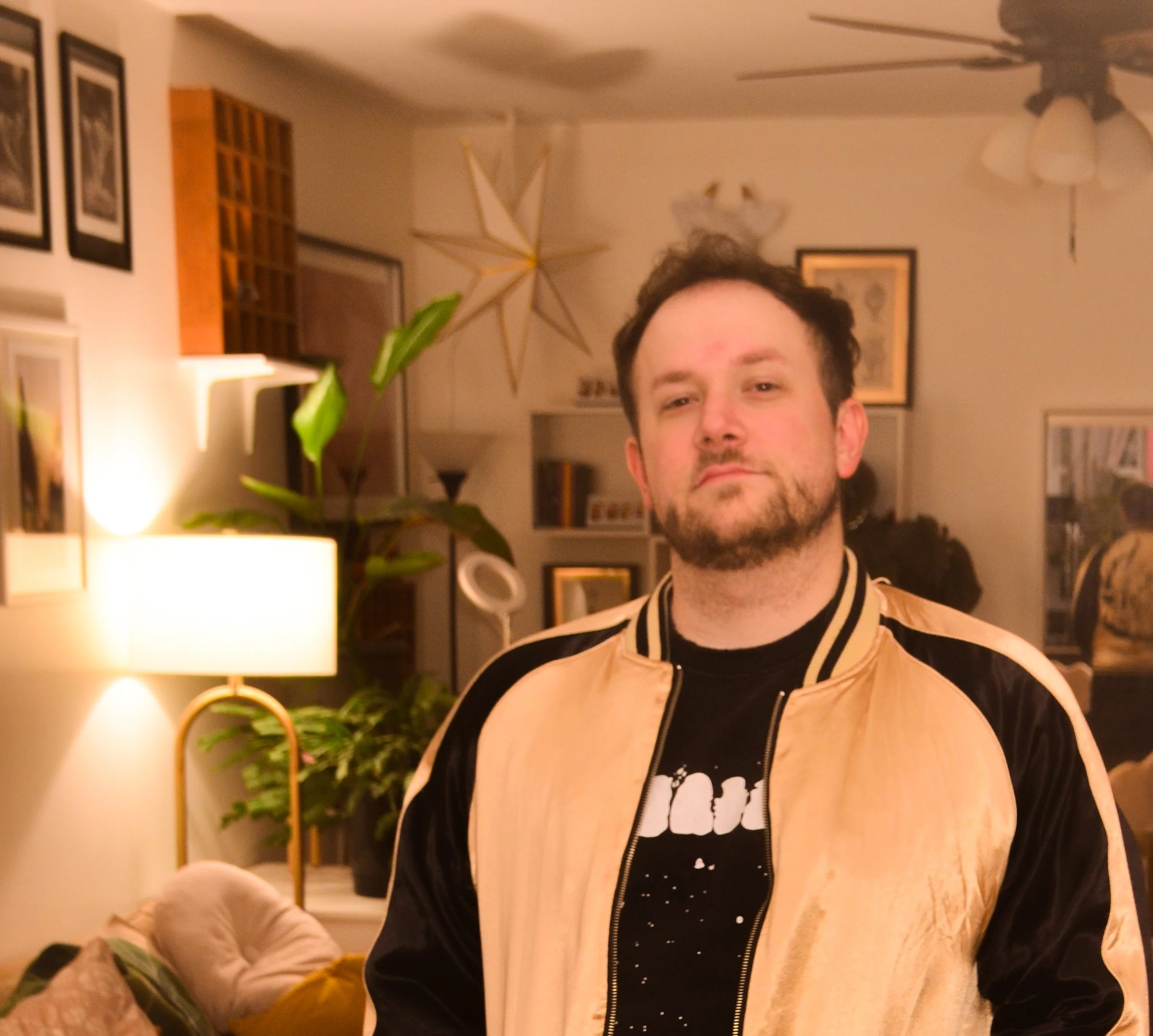 A picture of Kyle, the freelance publicist, standing in a room looking happy. They have on a gold and black jacket with a black shirt under it with teeth on it.