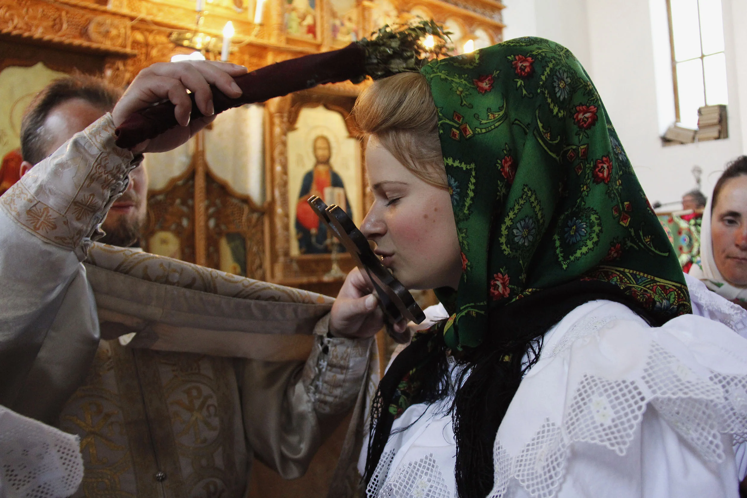  The priest blesses Marii by tapping her atop the head with a bouquet of dried basil soaked in holy water as she kisses the cross he holds in his other hand. 