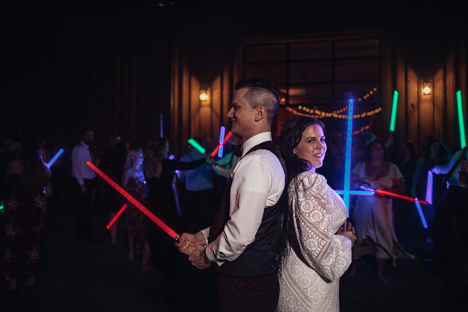 A bride and groom stand back to back with light sabers after a star wars send off.