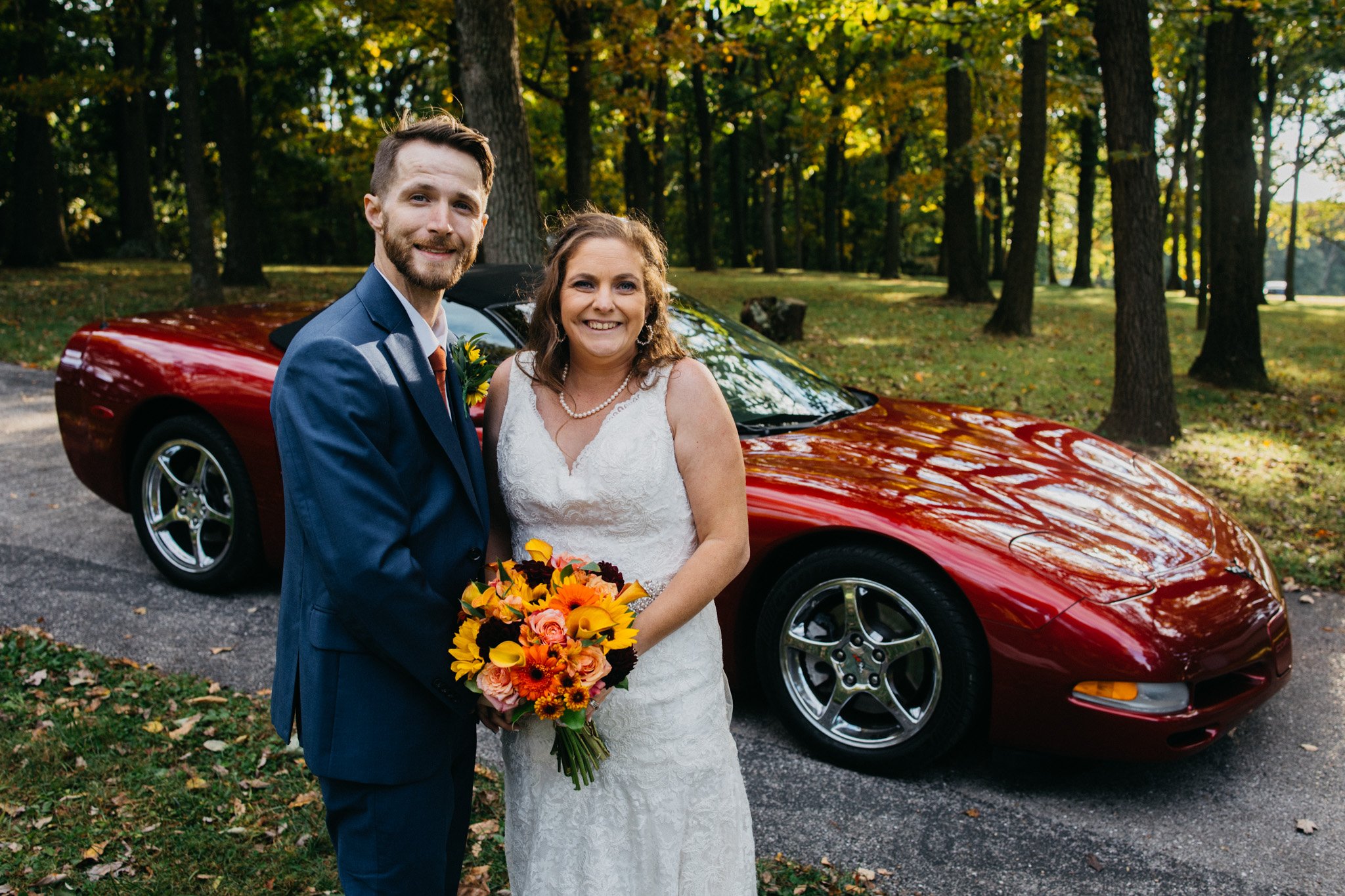 A bride and a groom pose in front of a red corvette.