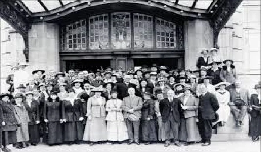 A large group of people, mostly women, are gathered on the steps outside a building with an ornate glass and metal awning. The photo appears to be vintage, possibly early 20th century.