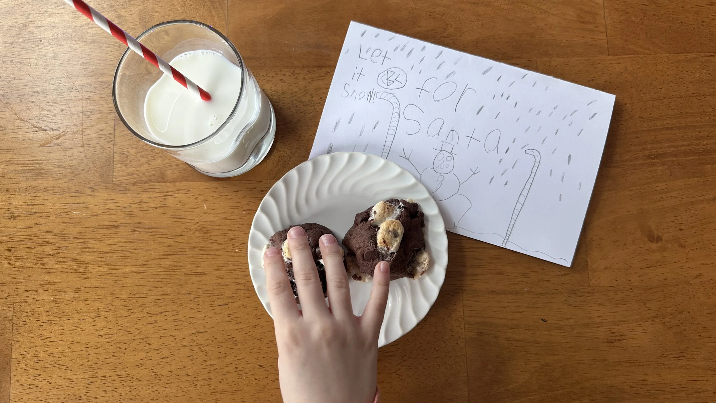 A child’s hand reaching for brownie cookies with marshmallows on a white plate next to a glass of milk and a handwritten note for Santa.