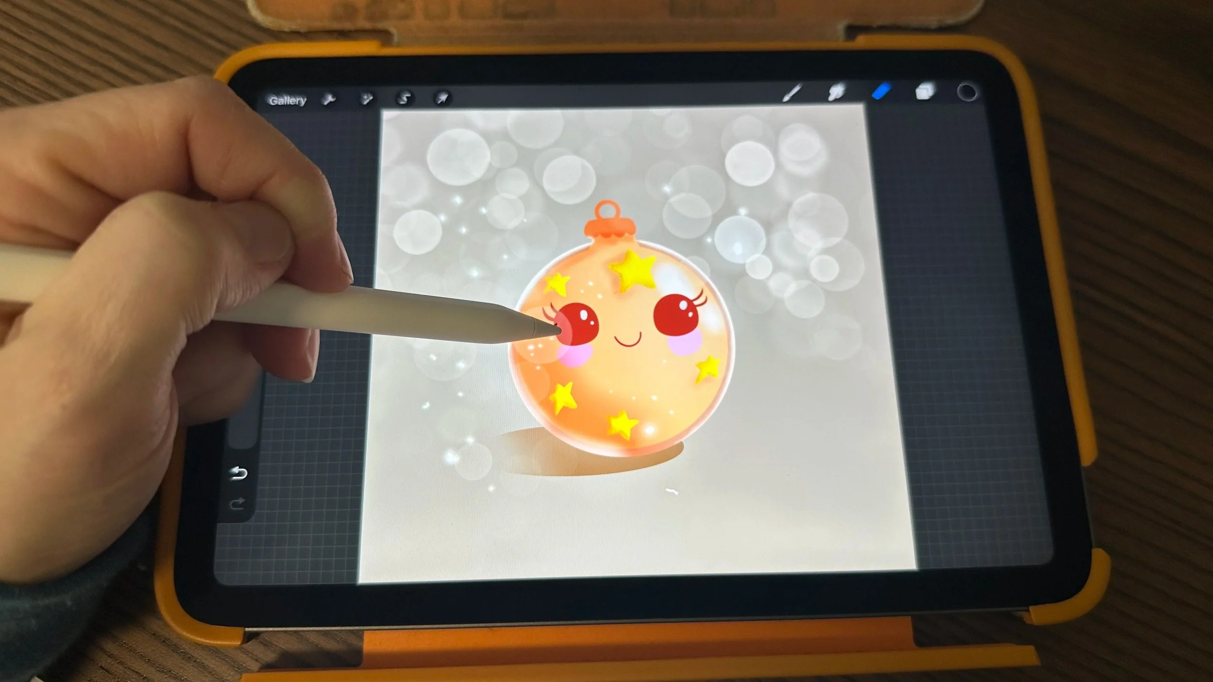 Hand holding an Apple Pencil over an iPad in Procreate, drawing a cute ornament character on a sparkly bokeh background.
