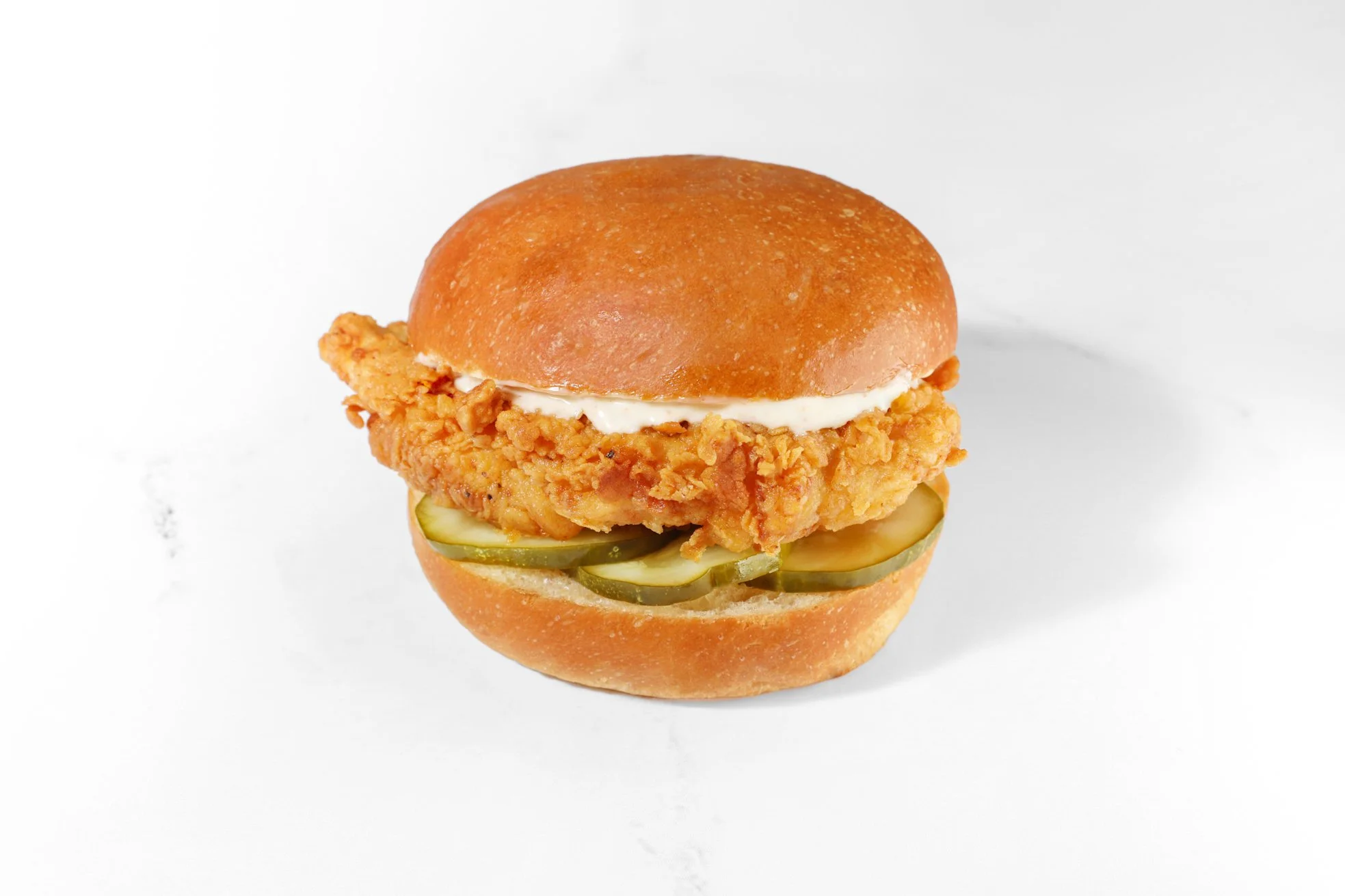Commercial food photography of a crispy fried chicken sandwich topped with pickles on a toasted bun.