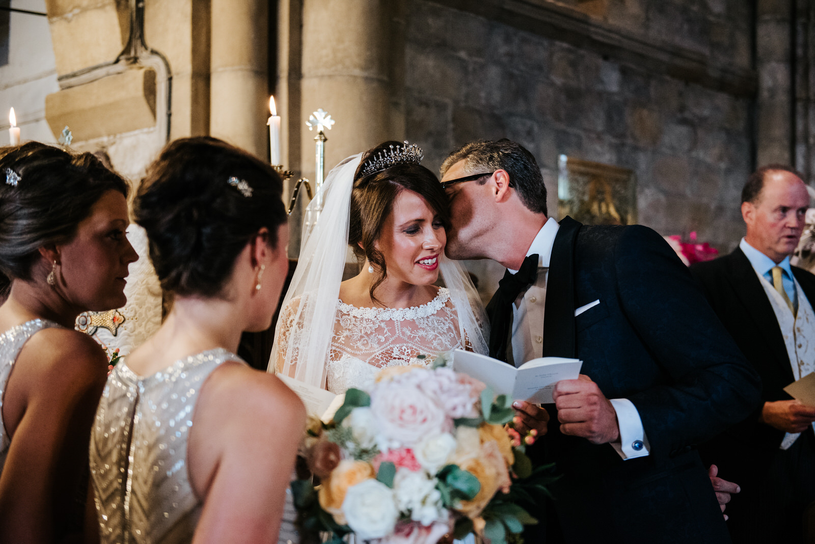 Groom kisses bride on the cheek inside church as readings take p