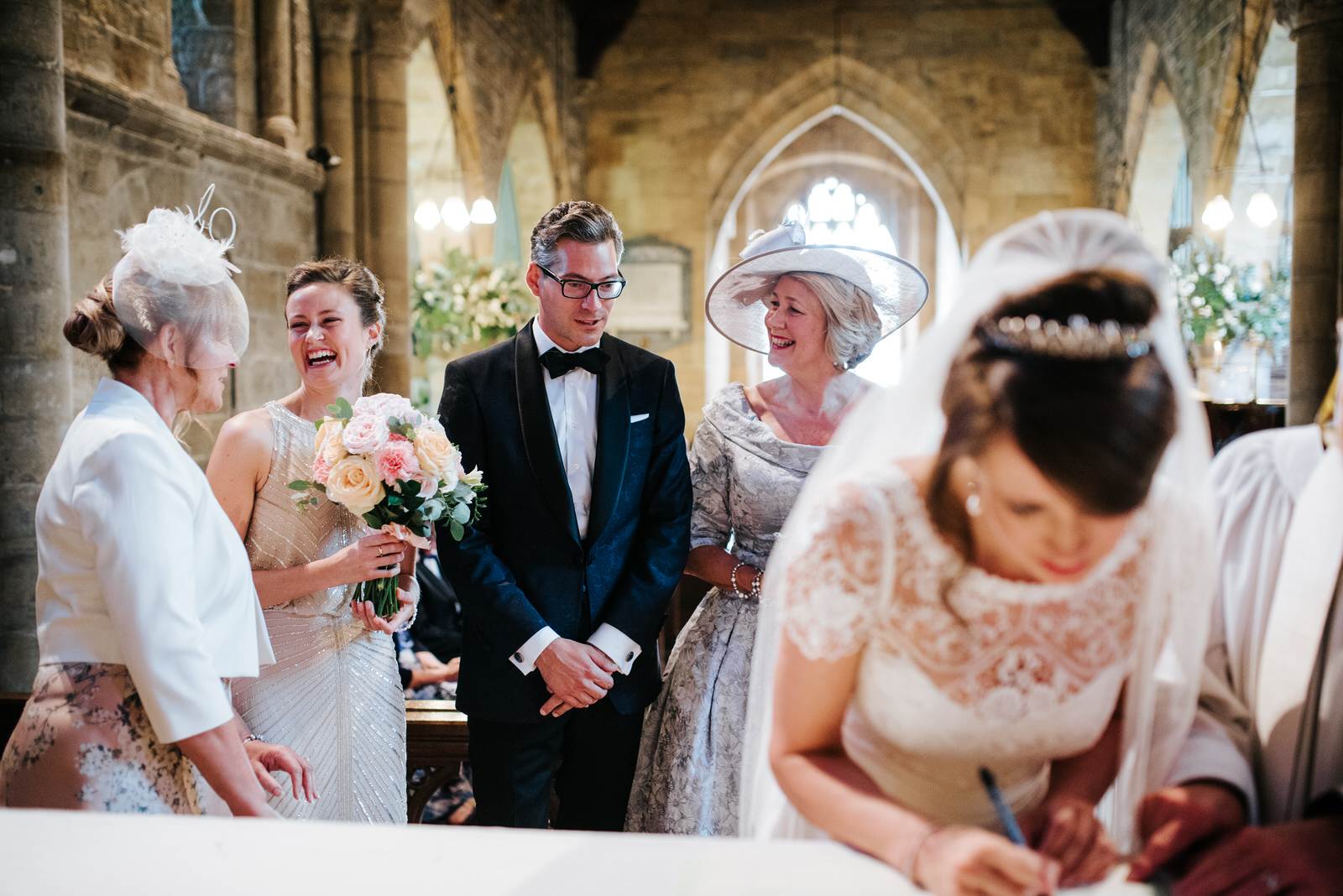 Bride signs the register while Groom and relatives laugh from be