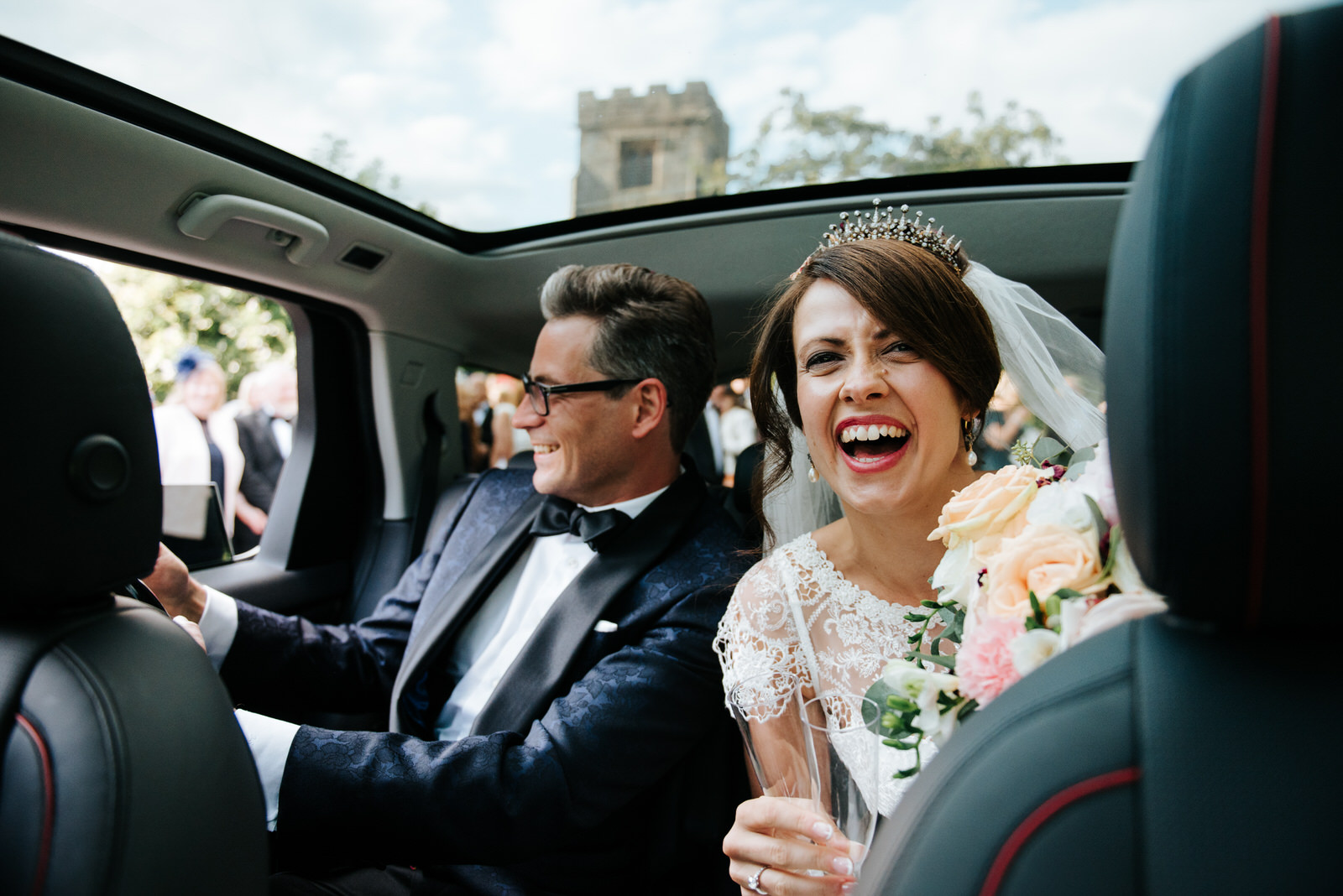 Bride and Groom sit inside car and open bottle of Champagne whil