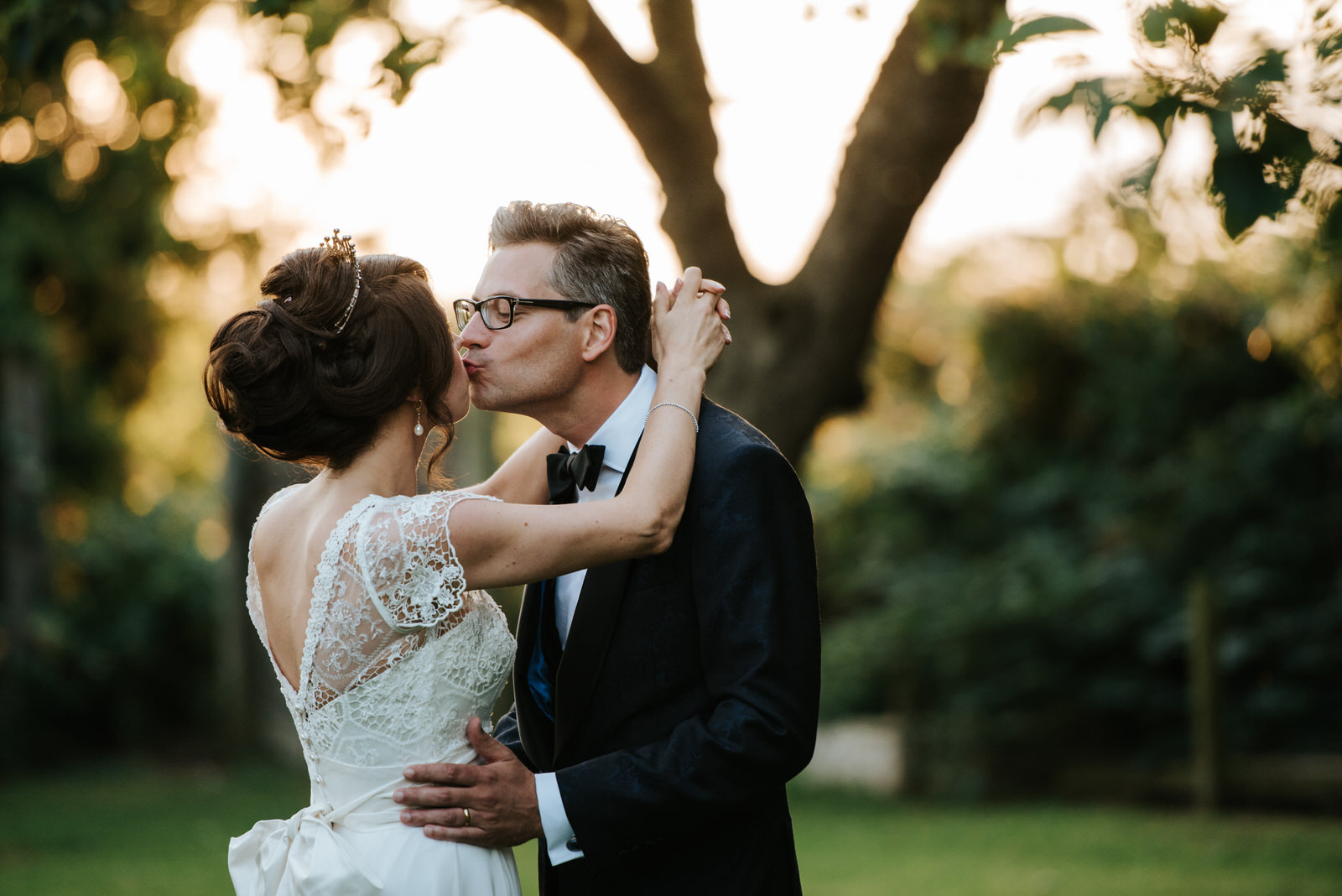 Bride and groom kiss in front of tree during portraits as sun se