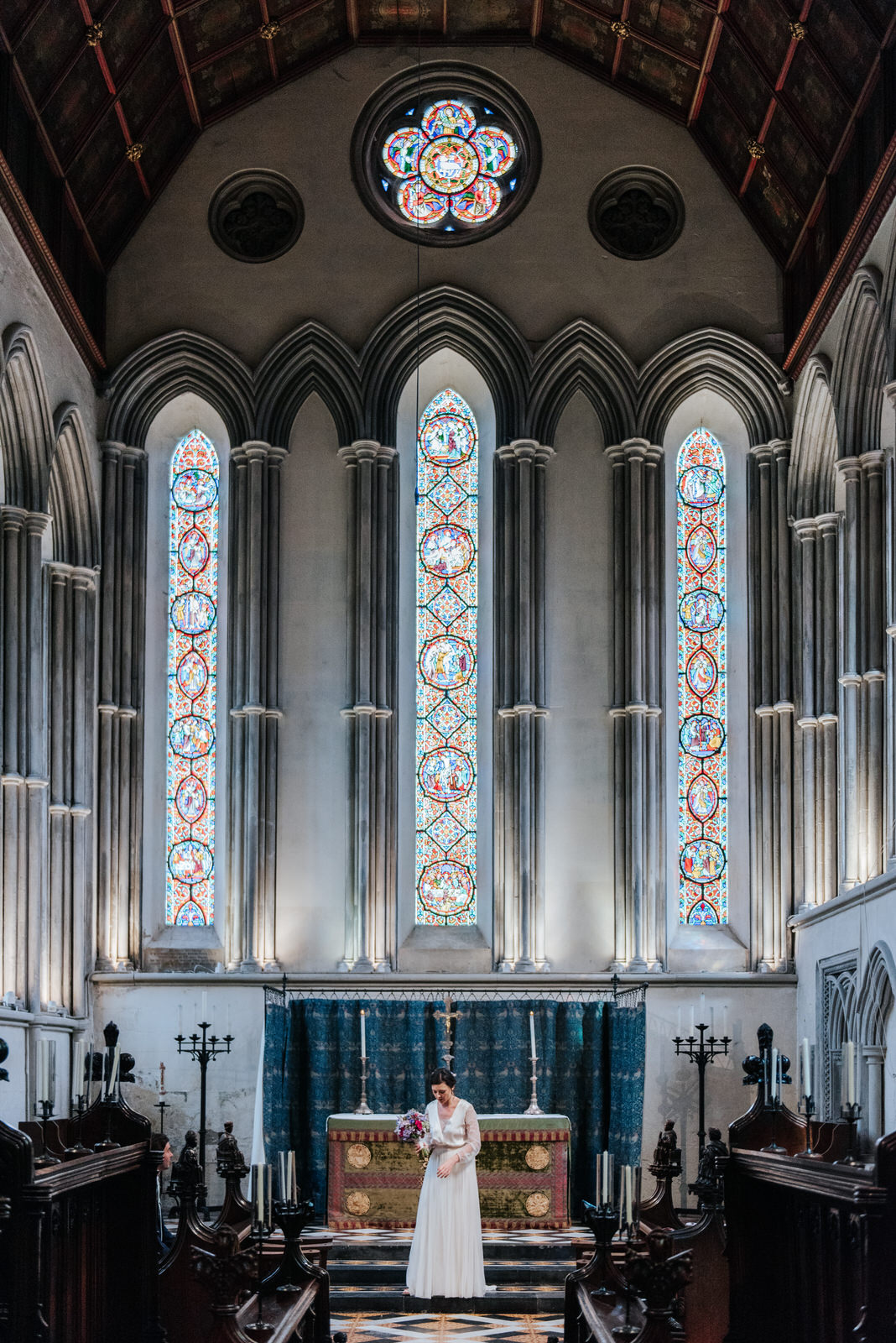 Bride stands under stained glass in Jesus College Chapel in Camb