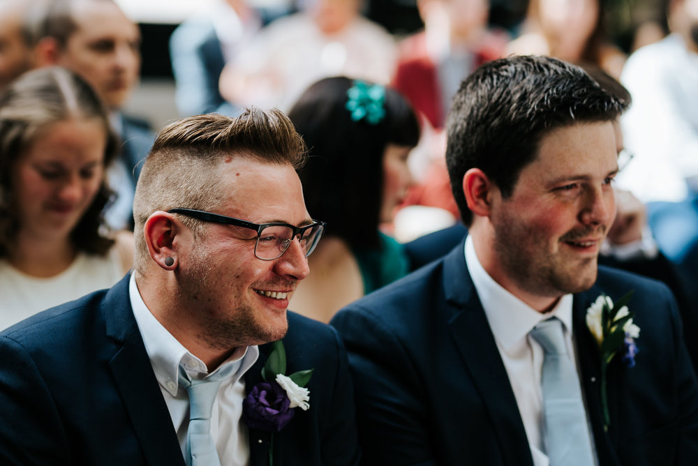 Two groomsmen smile and look joyfully towards bride and groom