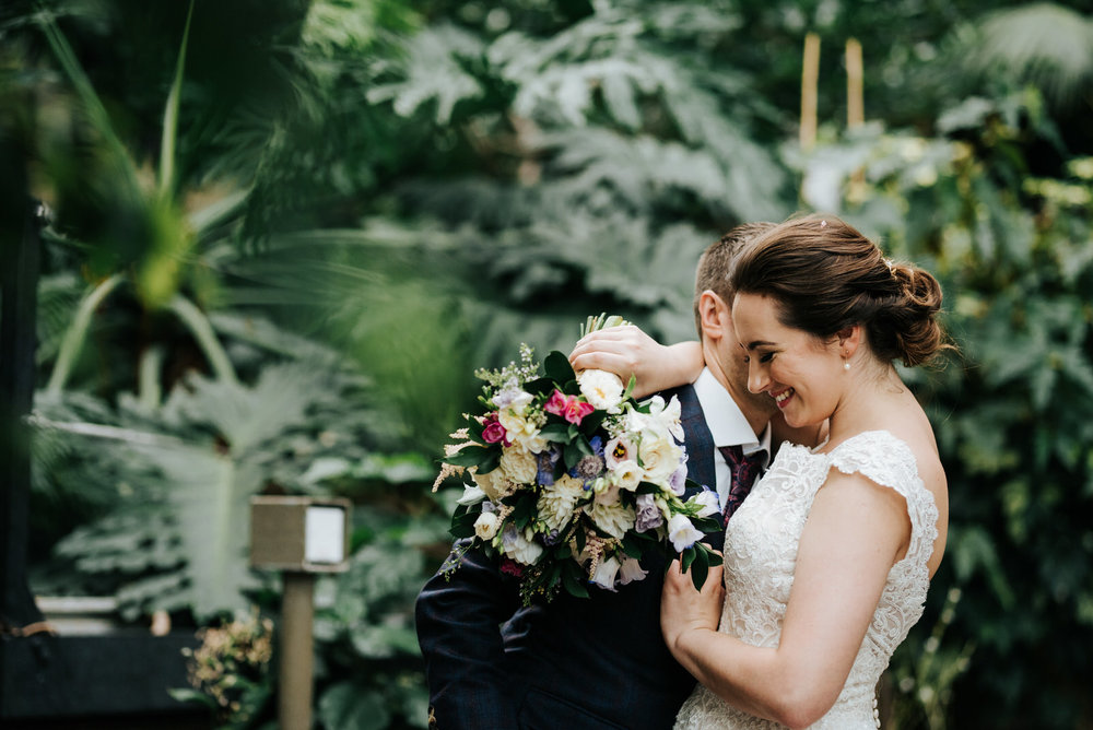 Groom whispers into brides ear as she smiles while hugging him w