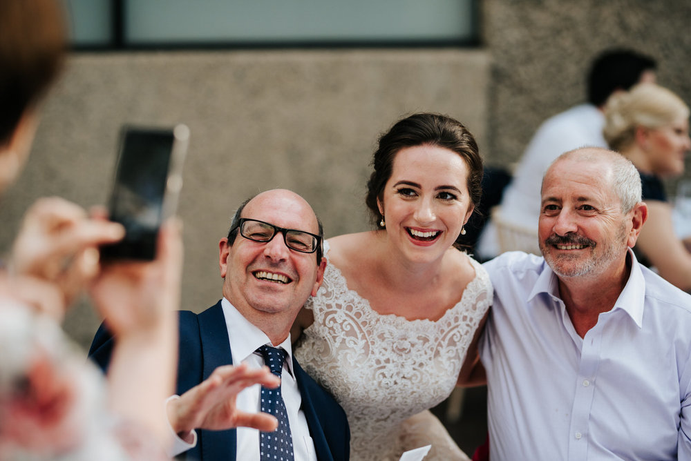  Bride and two family members smile into phone camera as they are photographed 