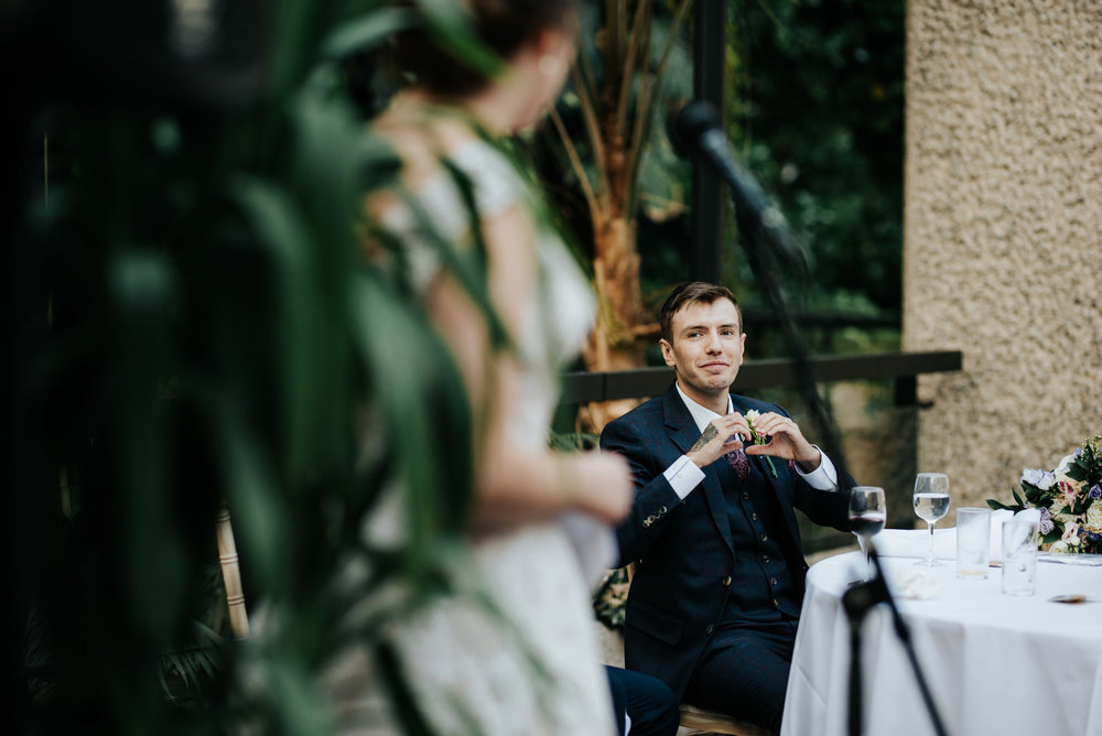 Groom looks towards bride with a heart formed with his hands aft