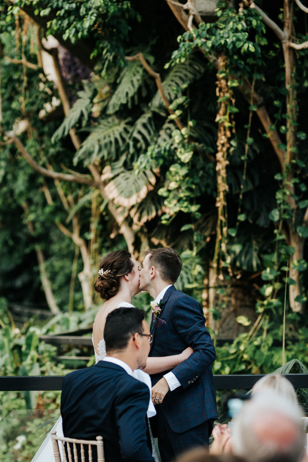 Groom kisses bride on the cheek while gorgeous greenery can be s