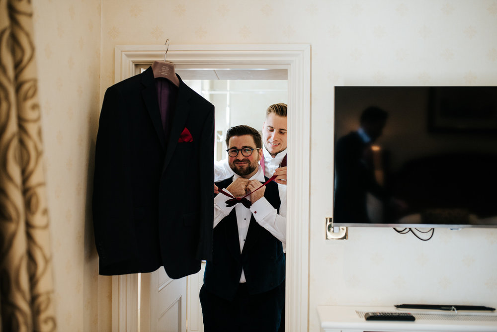 Groom stands by door as groomsman helps him put on his tie