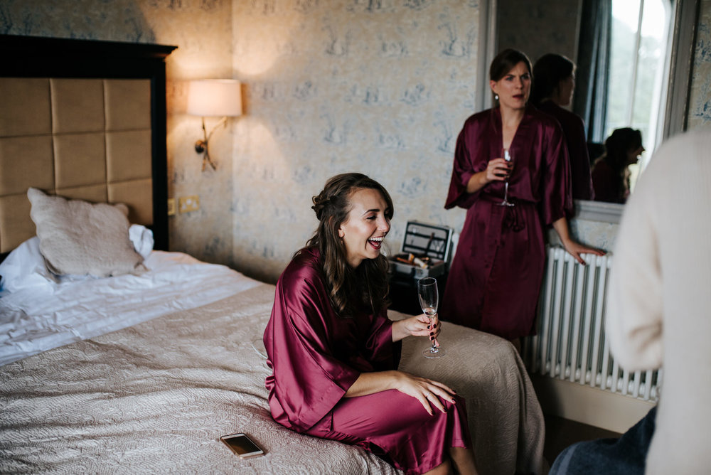 Bride's sister sits on bed and smiles while holding glass of prosecco