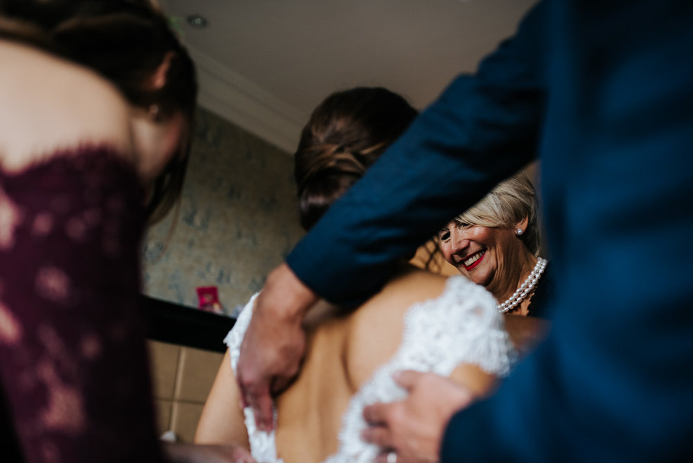 Bride's mum smiles as she helps bride put finishing touches on wedding dress