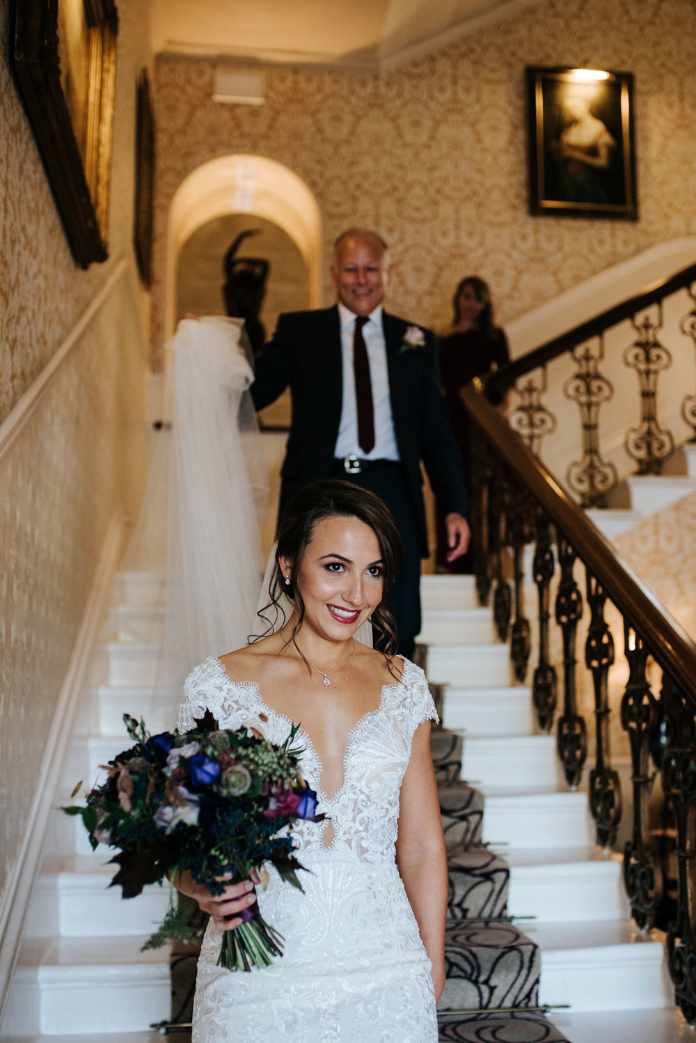 Bride walks down hotel stairs at The Petersham as step-dad holds her veil behind her
