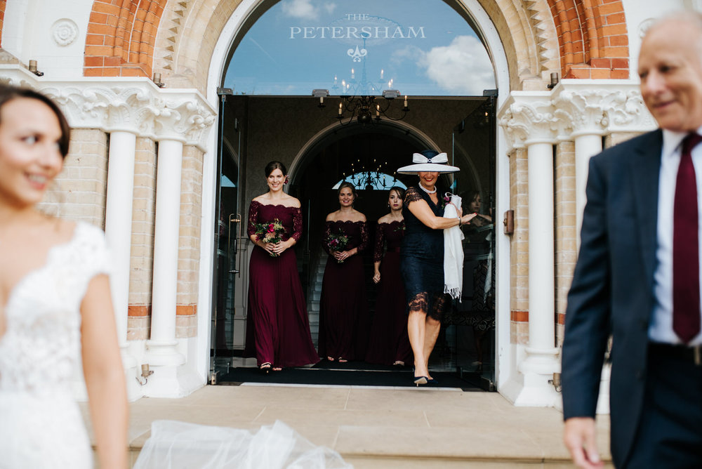 Bride and step-dad walk out of The Petersham as mum and bridesmaids smile from behind
