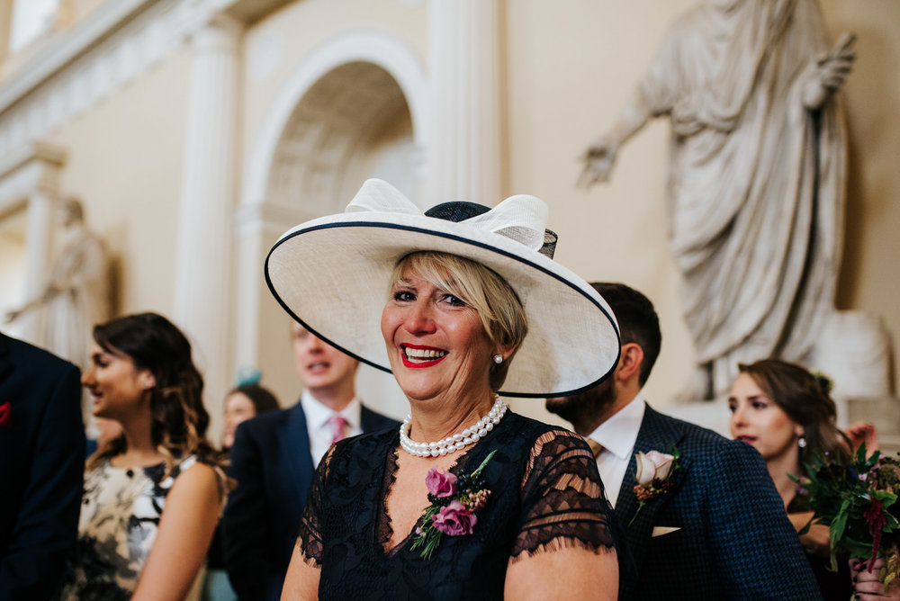 Bride's mum wears stunning  hat and smiles at camera as bride comes down aisle