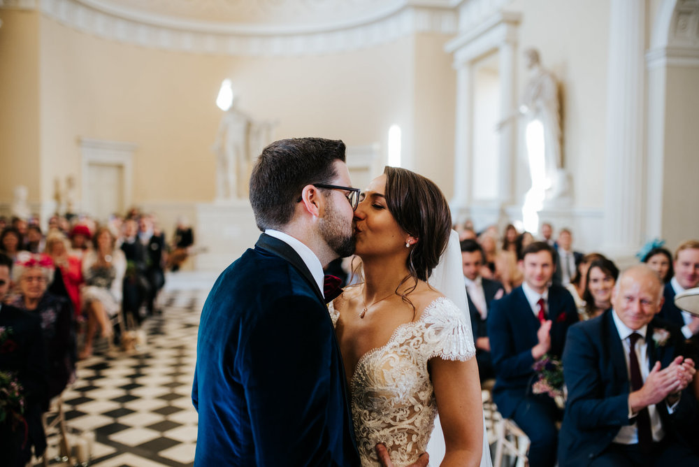 Bride and groom share their first kiss during ceremony at Syon House as guests look from back