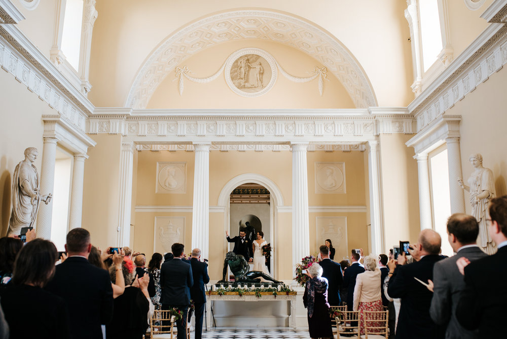 Bride and groom walk out of door flanked by stunning arches and statues as they are announced as husband and wife Syon House