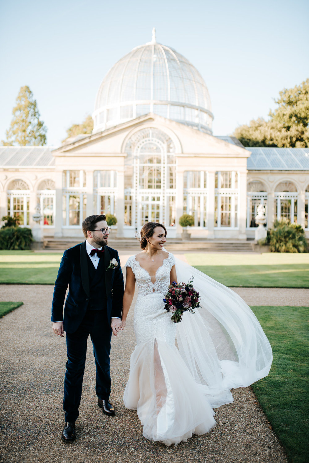 Bride and groom walk towards camera as great conservatory building towers from behind them