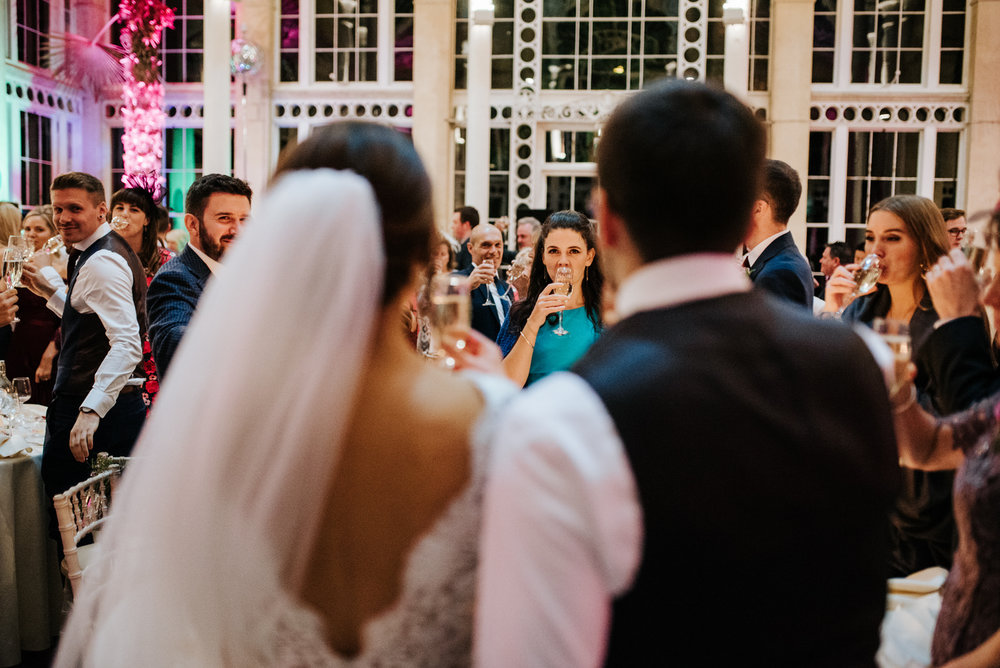 Bride and groom stand centre, out of focus, as guests toast behind them