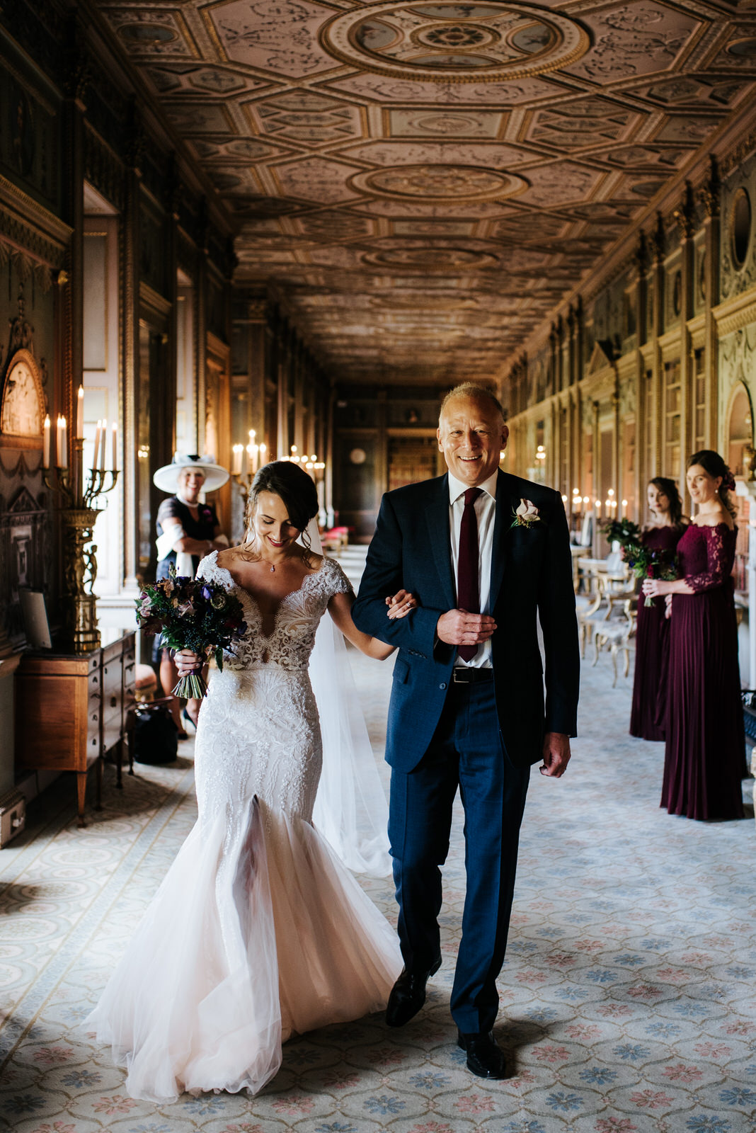 Bride and step-dad walk towards the aisle as mum and bridesmaids look joyfully from behind