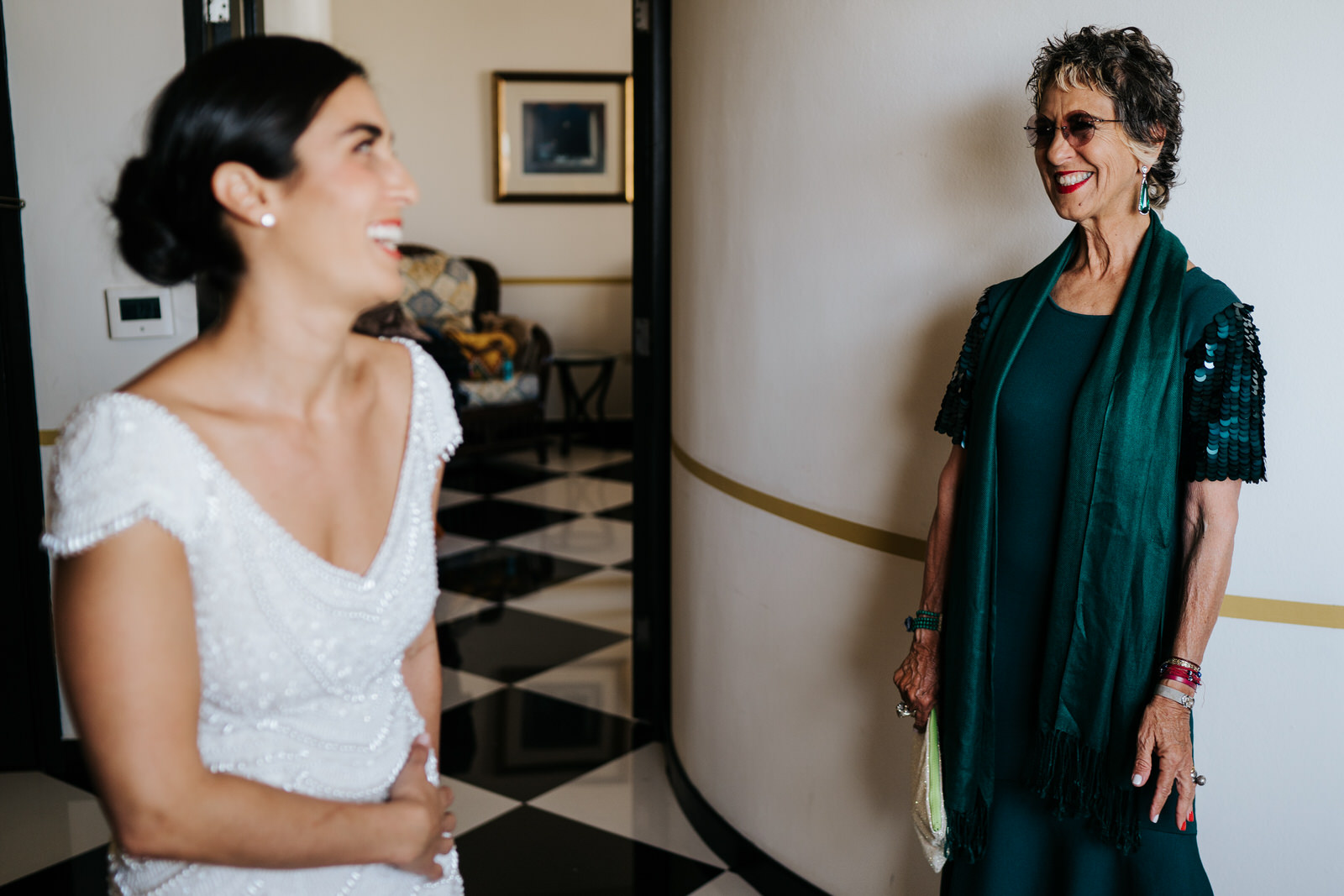 Bride, in front, out of focus as her aunt looks and smiles to her while she is getting ready for the wedding in hotel room