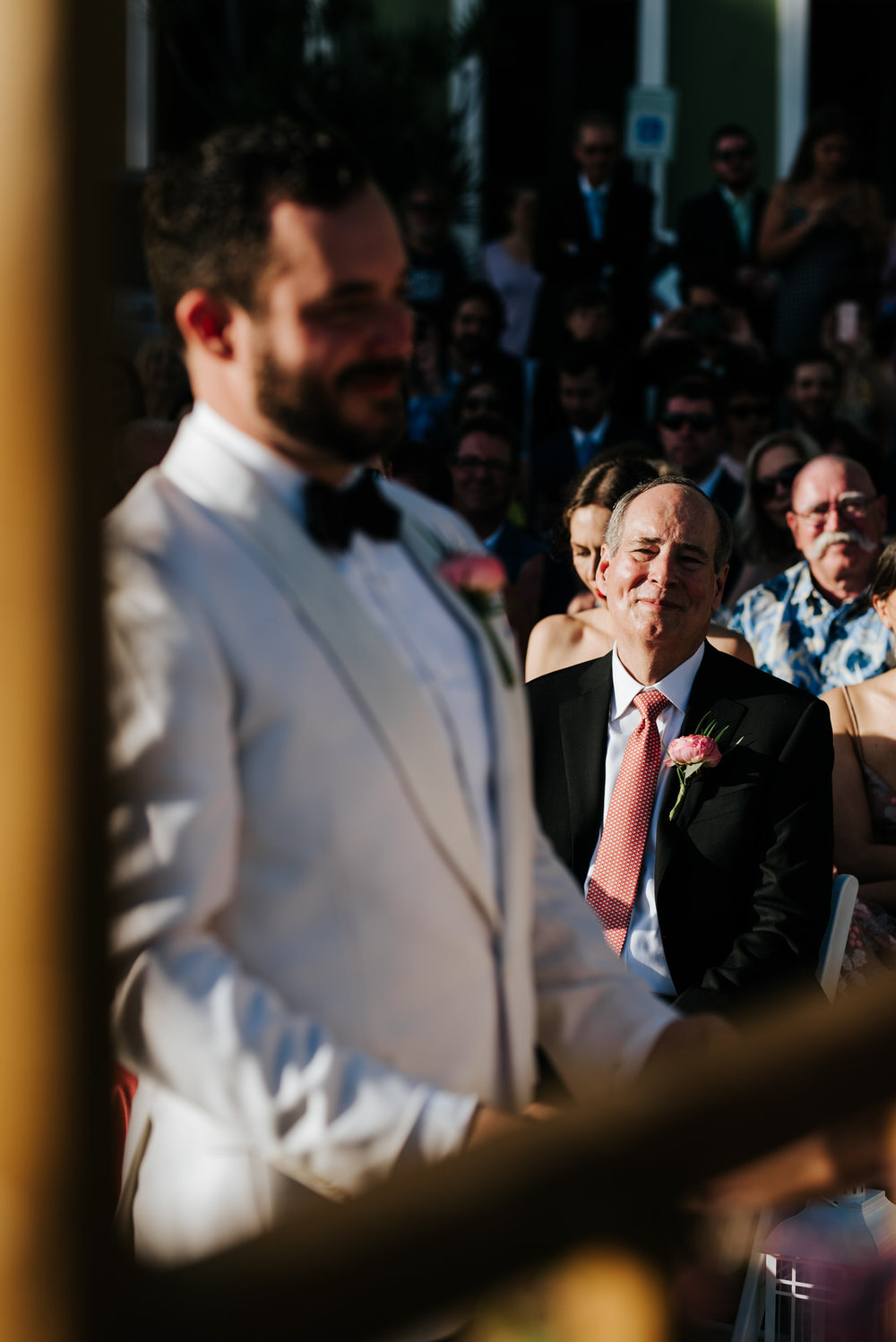  Groom stands, out of focus, at the front of the aisle as his father smiles towards him from the background 