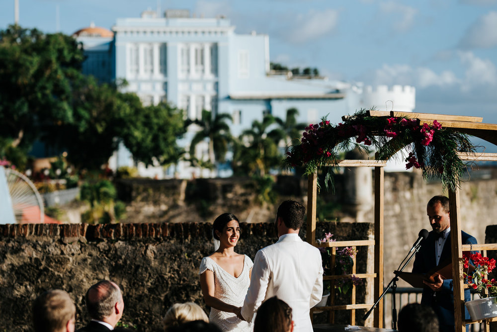  Wide photograph of where the wedding ceremony took place at La Rogativa in Old San Juan, Puerto Rico with the bride and groom holding hands and smiling at the front and La Fortaleza in the back 