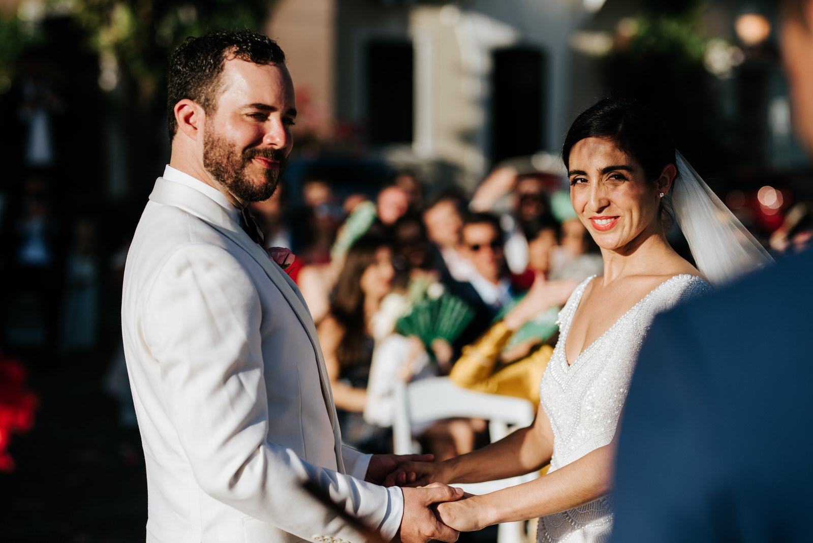 Close-up photograph of bride and groom holding hands and smiling at their officiant as he conducts the wedding ceremony