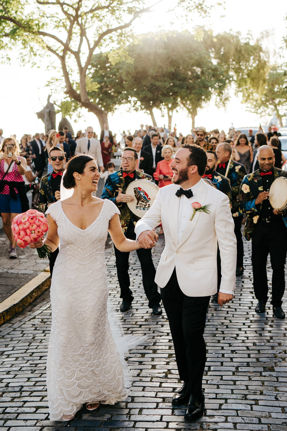  Bride and groom hold hands and smile at each other while the sun shines and musicians and guests follow behind them 